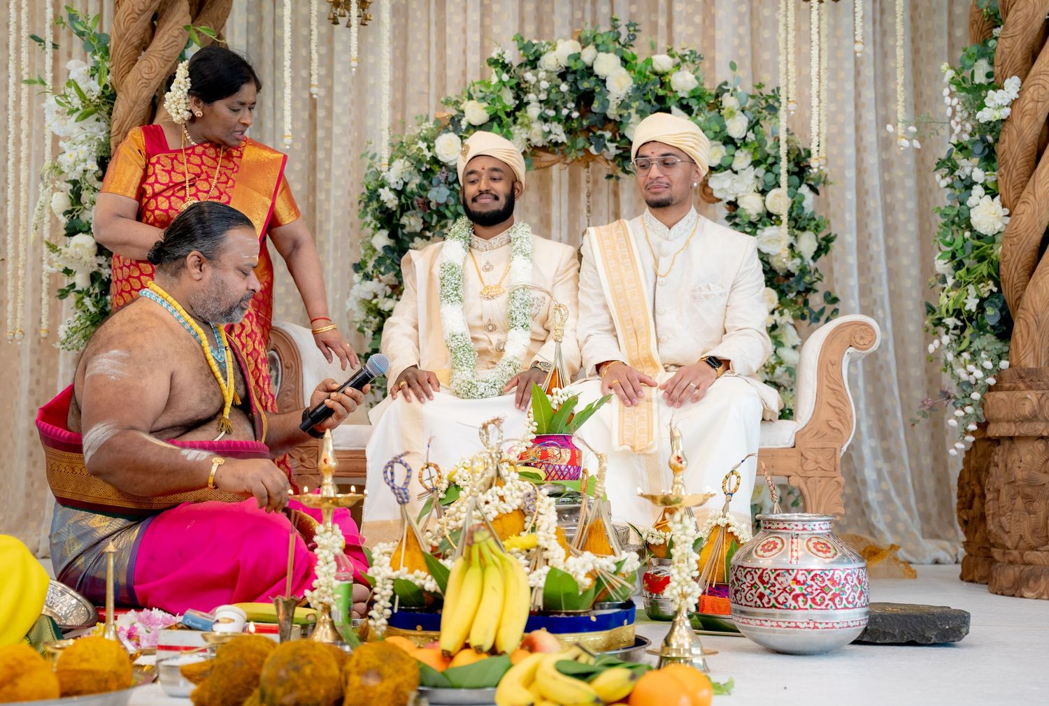 Two grooms in white attire sit at a Hindu wedding ceremony with a priest and floral backdrop.