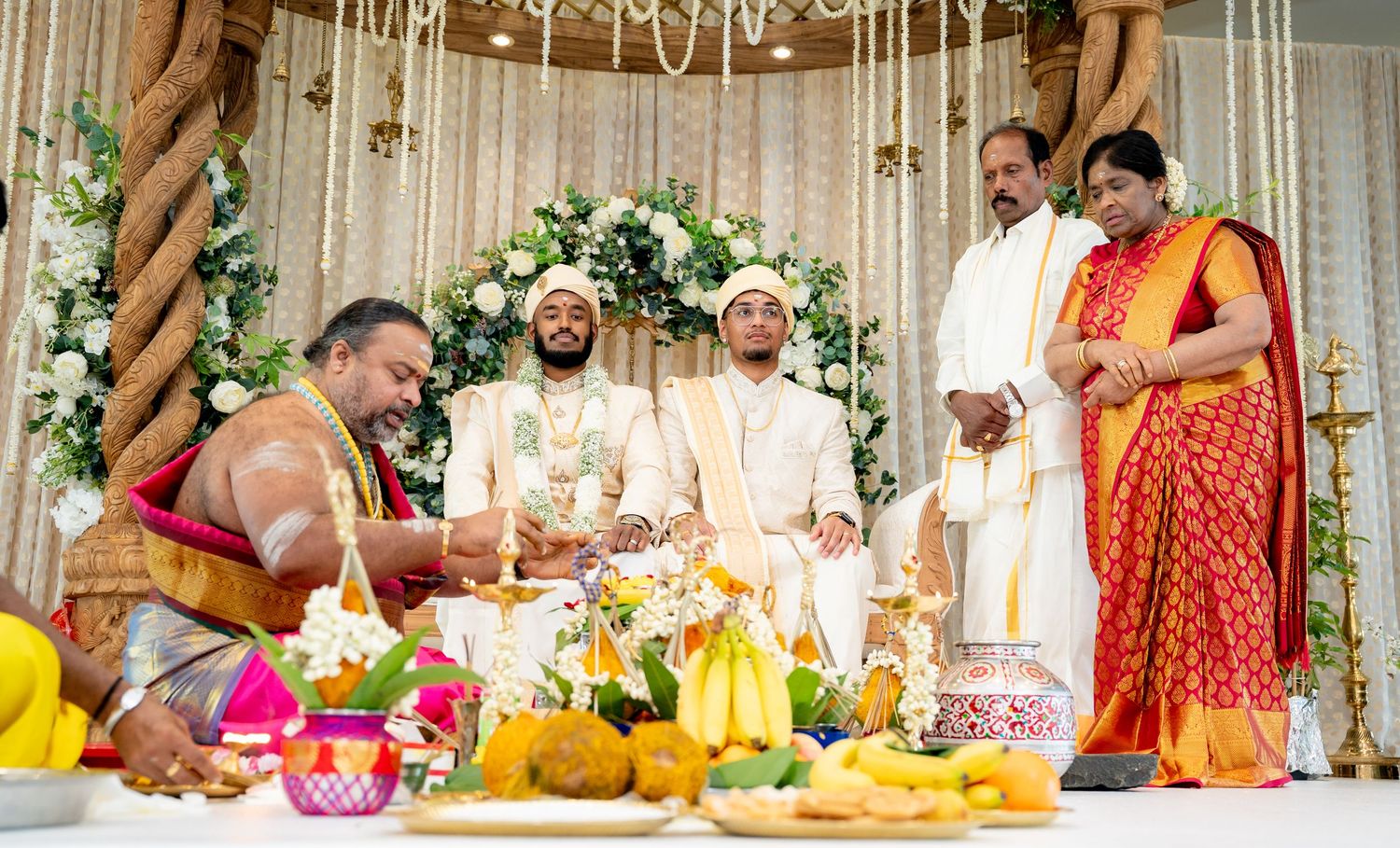 South Indian wedding ritual with priest performing ceremony for groom in white attire at floral decorated altar.