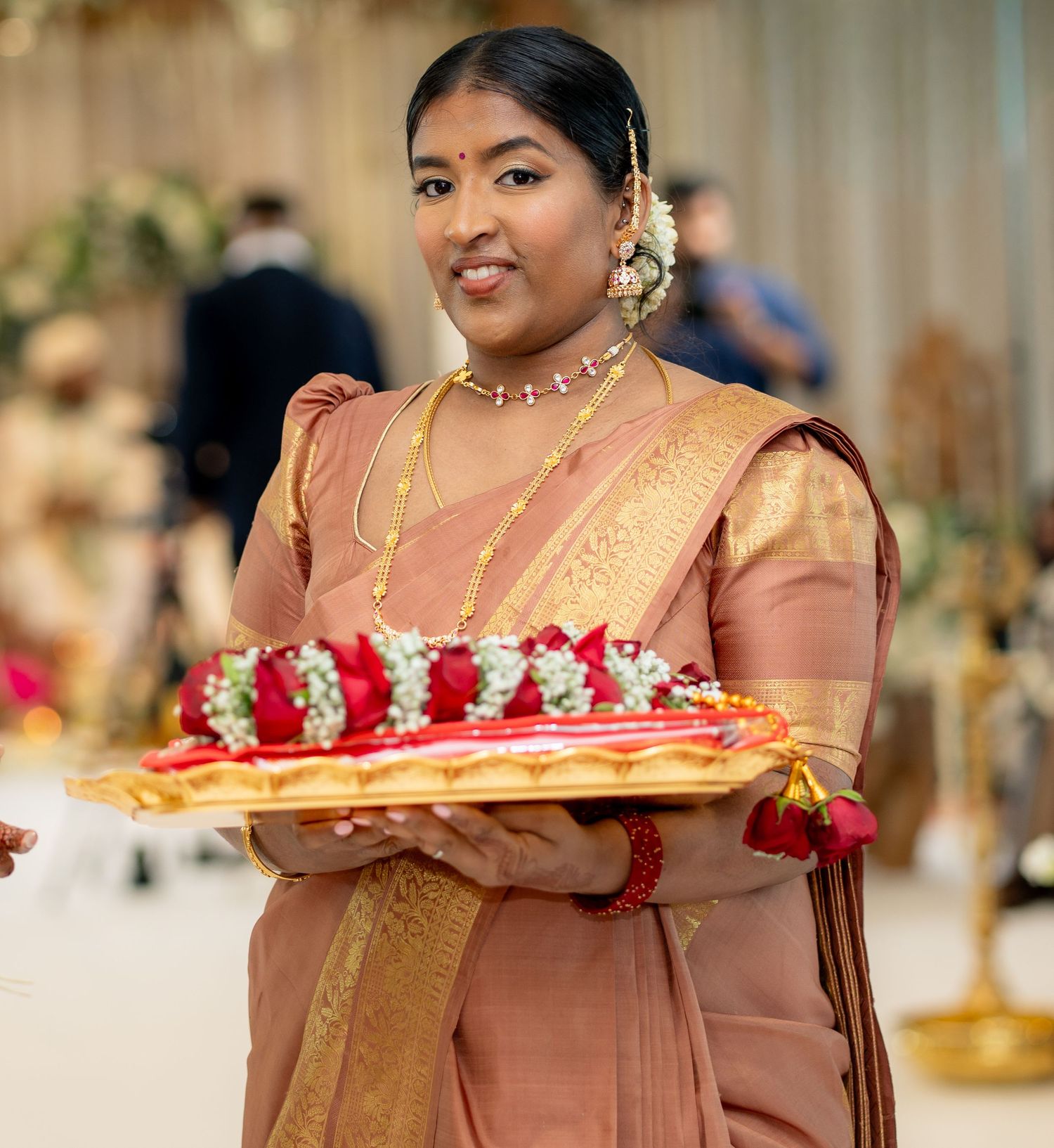Woman in rose gold silk saree holds tray of red and white flower garlands at Indian wedding ceremony.