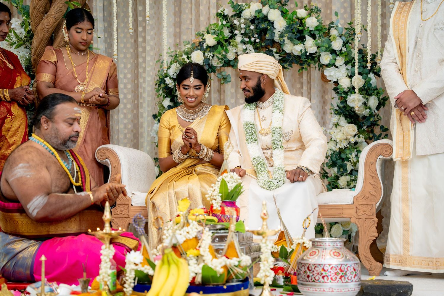 Traditional South Indian wedding ceremony with bride in gold saree and groom in white sherwani, exchanging vows with priest.