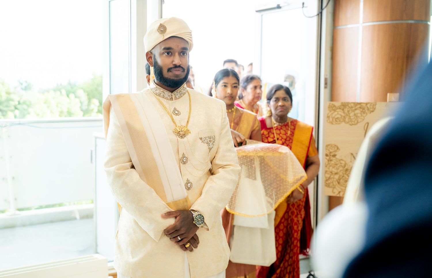 Groom in cream sherwani and golden turban walking into a traditional Hindu wedding ceremony with guests in background.