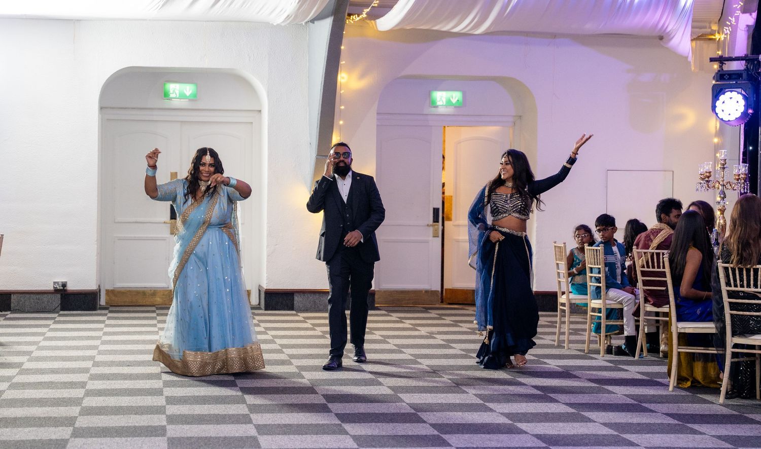 Two women in blue lehengas perform a dance entrance at an elegant Indian wedding reception hall.