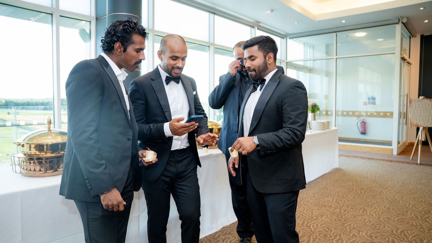 Three men in tuxedos gathered together looking at a smartphone at a formal event.
