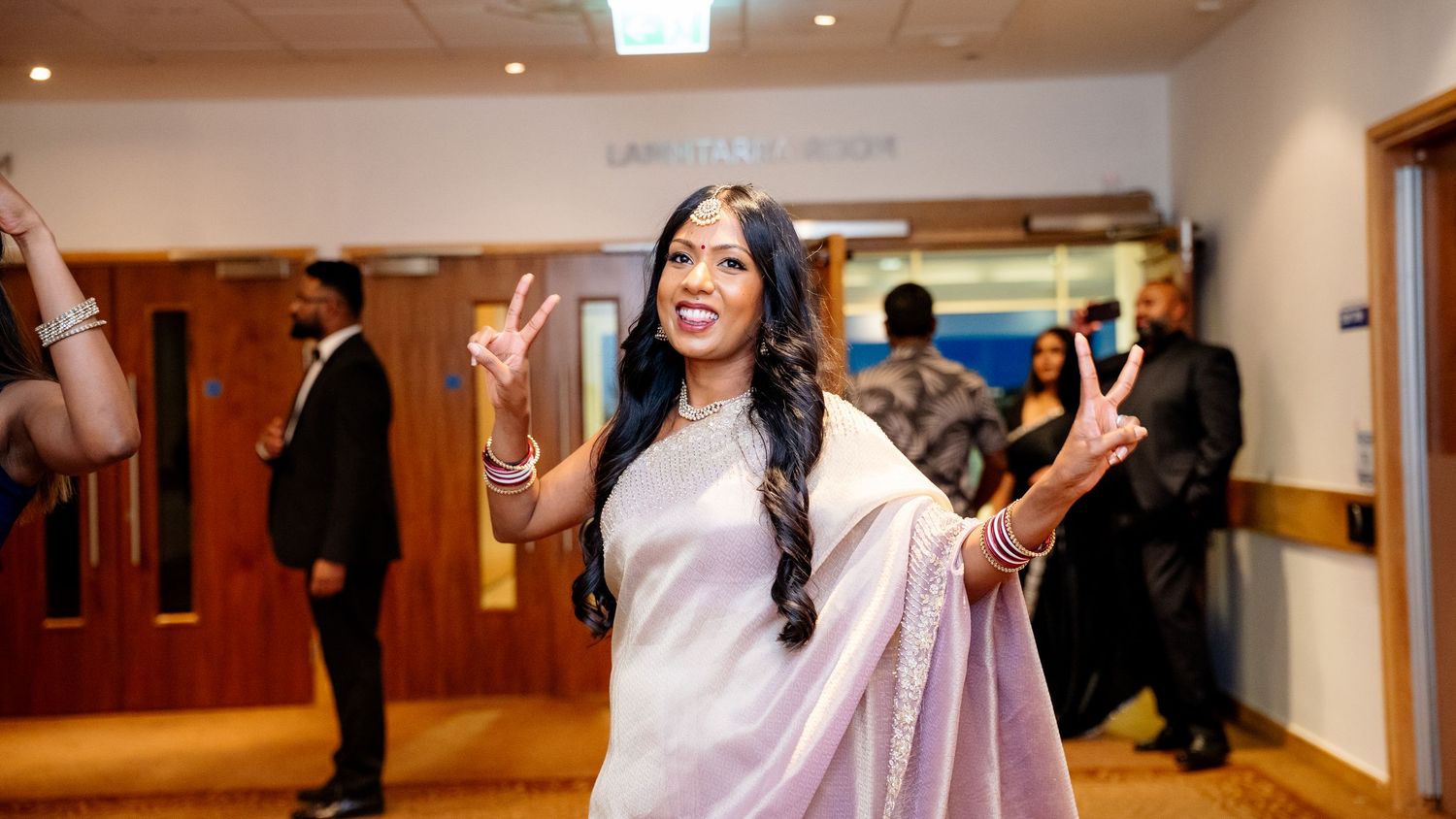 A woman in a lavender saree smiles and makes peace signs at an elegant indoor wedding reception.