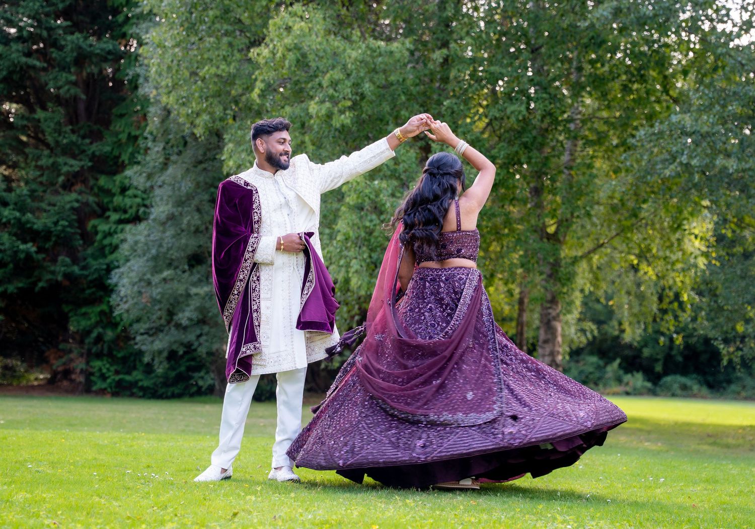Couple in purple and white Indian wedding attire dancing outdoors on green lawn with trees in background.
