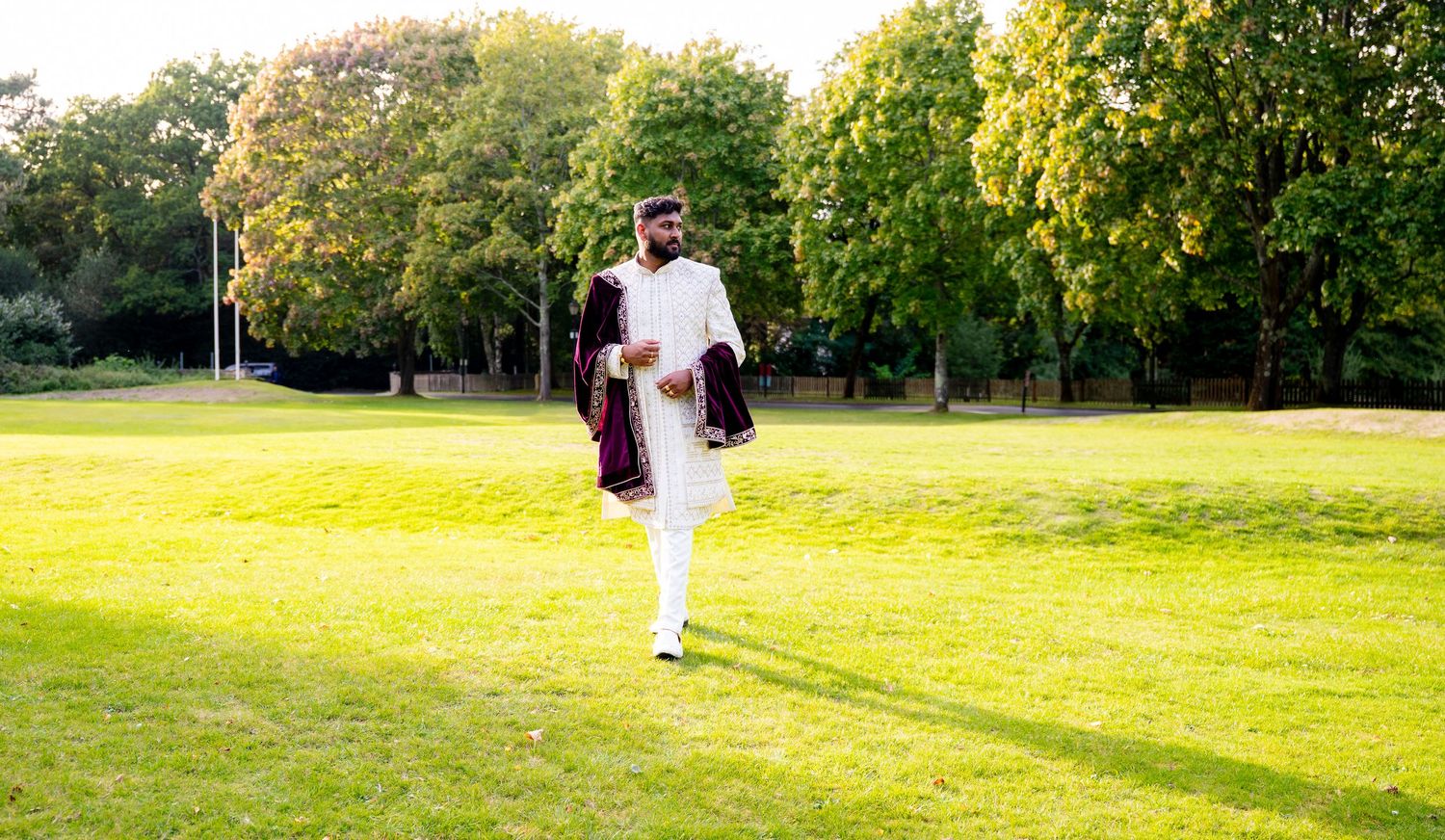 Indian groom in white sherwani and purple velvet shawl walking confidently across green park at outdoor wedding celebration.