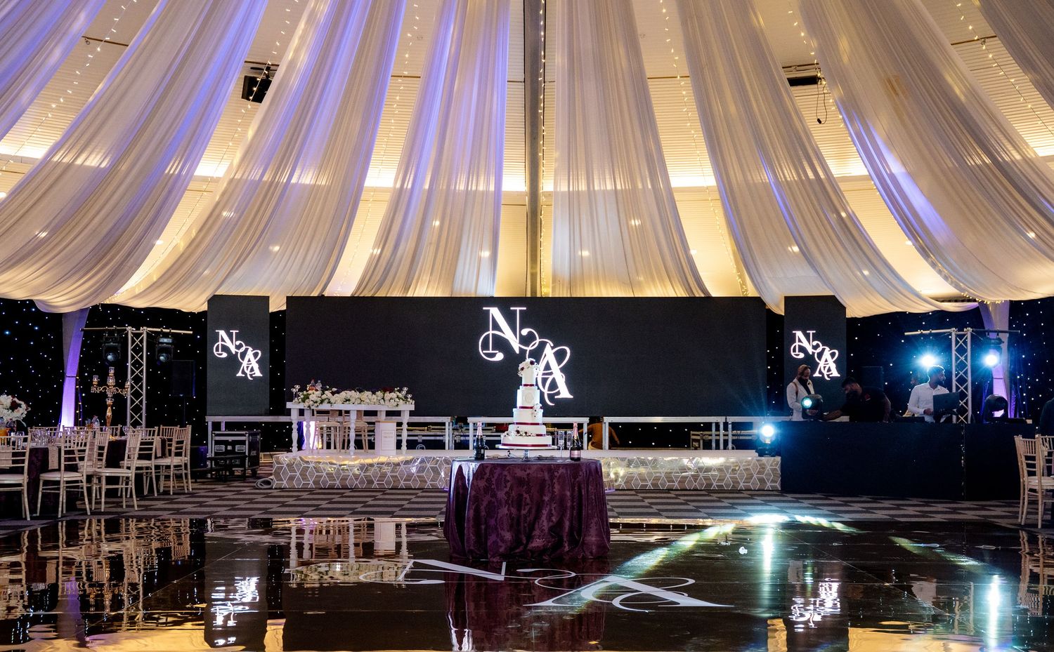 Elegant wedding reception venue with draped white ceiling, purple tablecloths, and illuminated monogram backdrop.