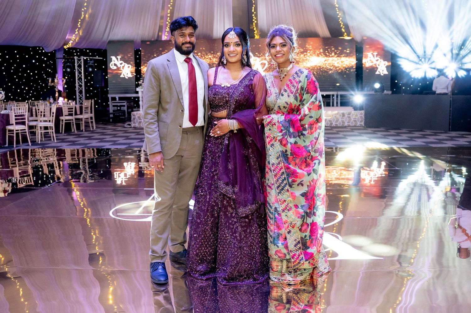 Three guests pose together on a decorated wedding dance floor, with the bride in a purple embroidered lehenga.