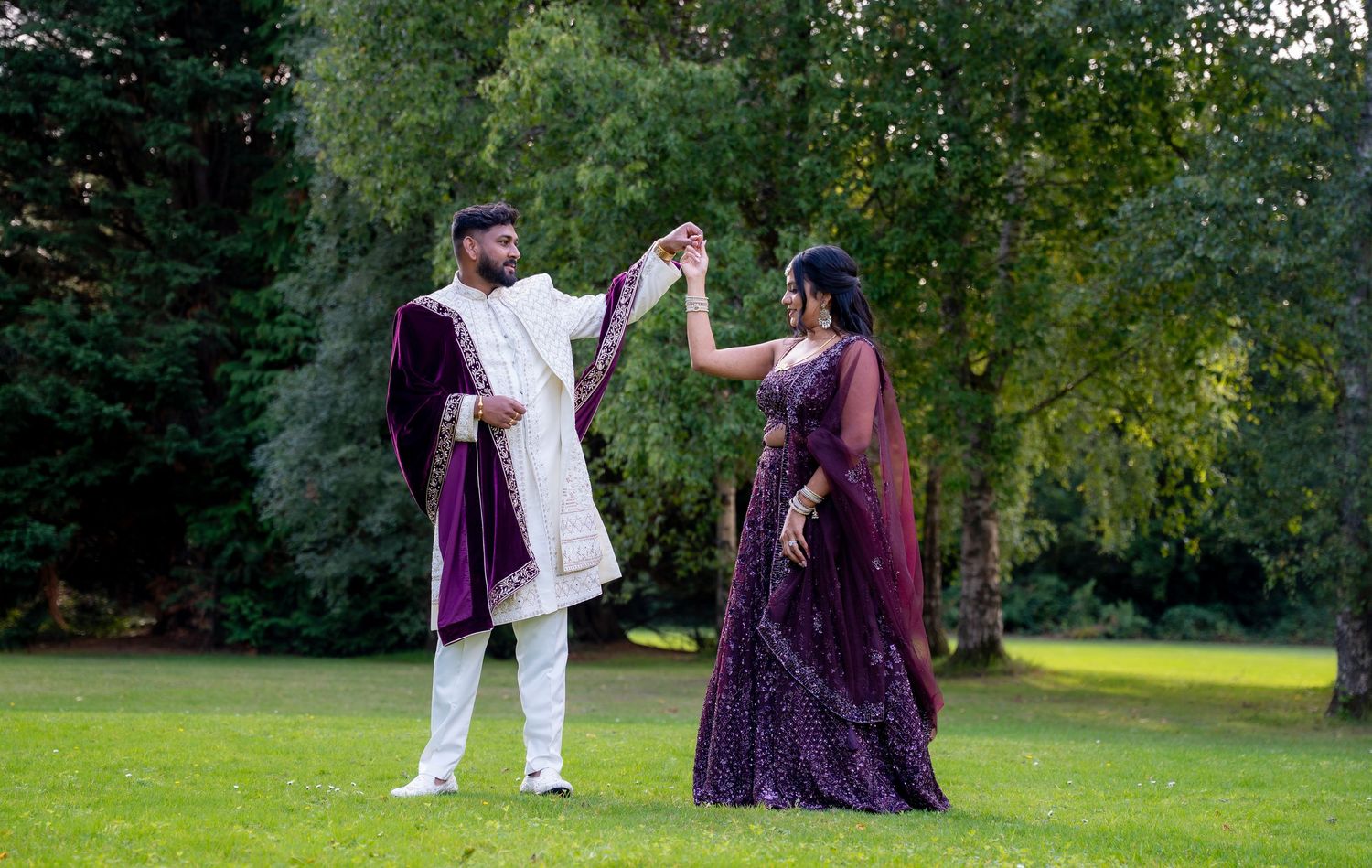 Couple in matching purple and white traditional South Asian wedding attire dancing outdoors in a lush green garden.