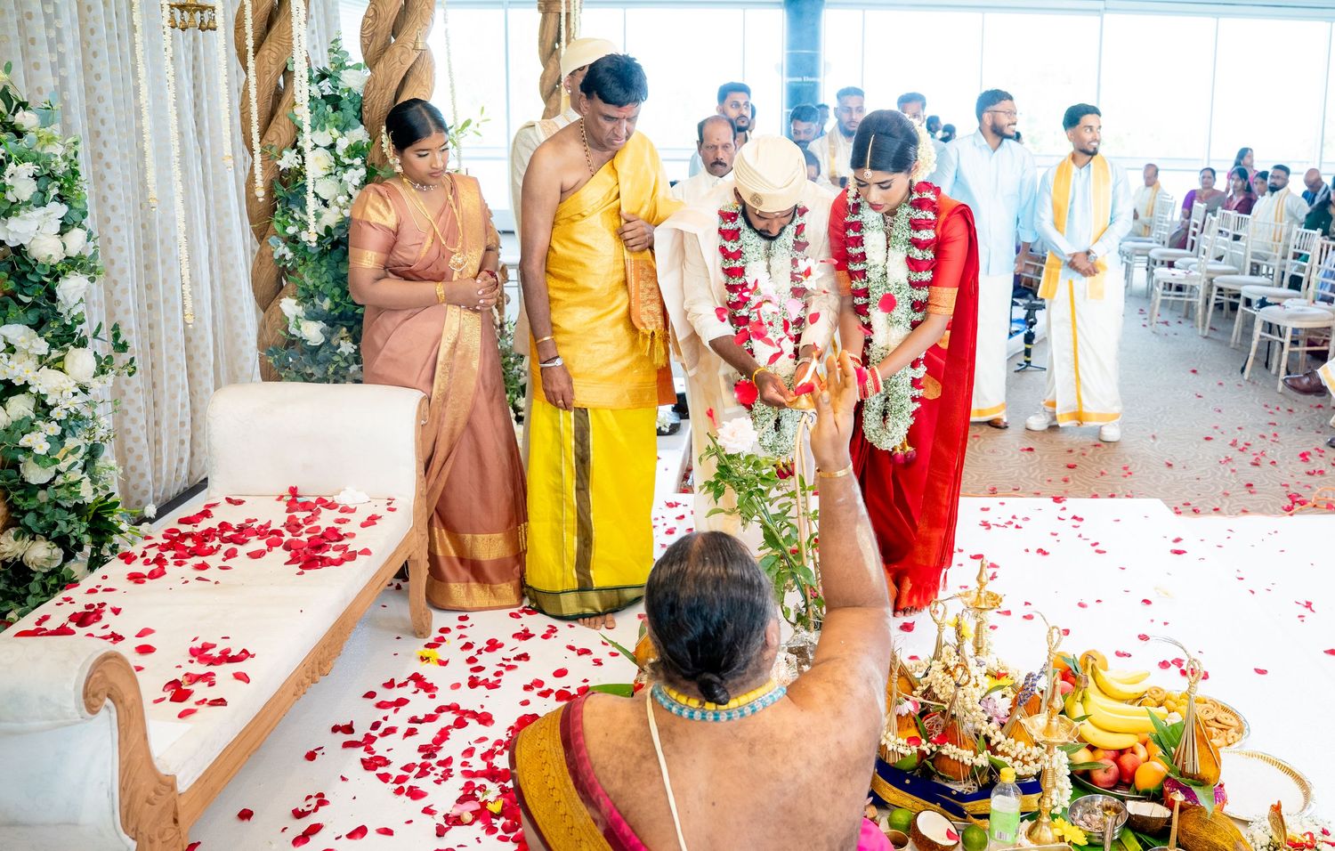 Priest performs Hindu wedding rituals with rose petals scattered on floor before bride and groom.