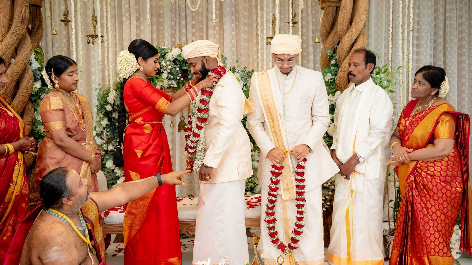 Traditional Hindu wedding garland exchange with bride in red saree and groom in white sherwani at floral mandap.