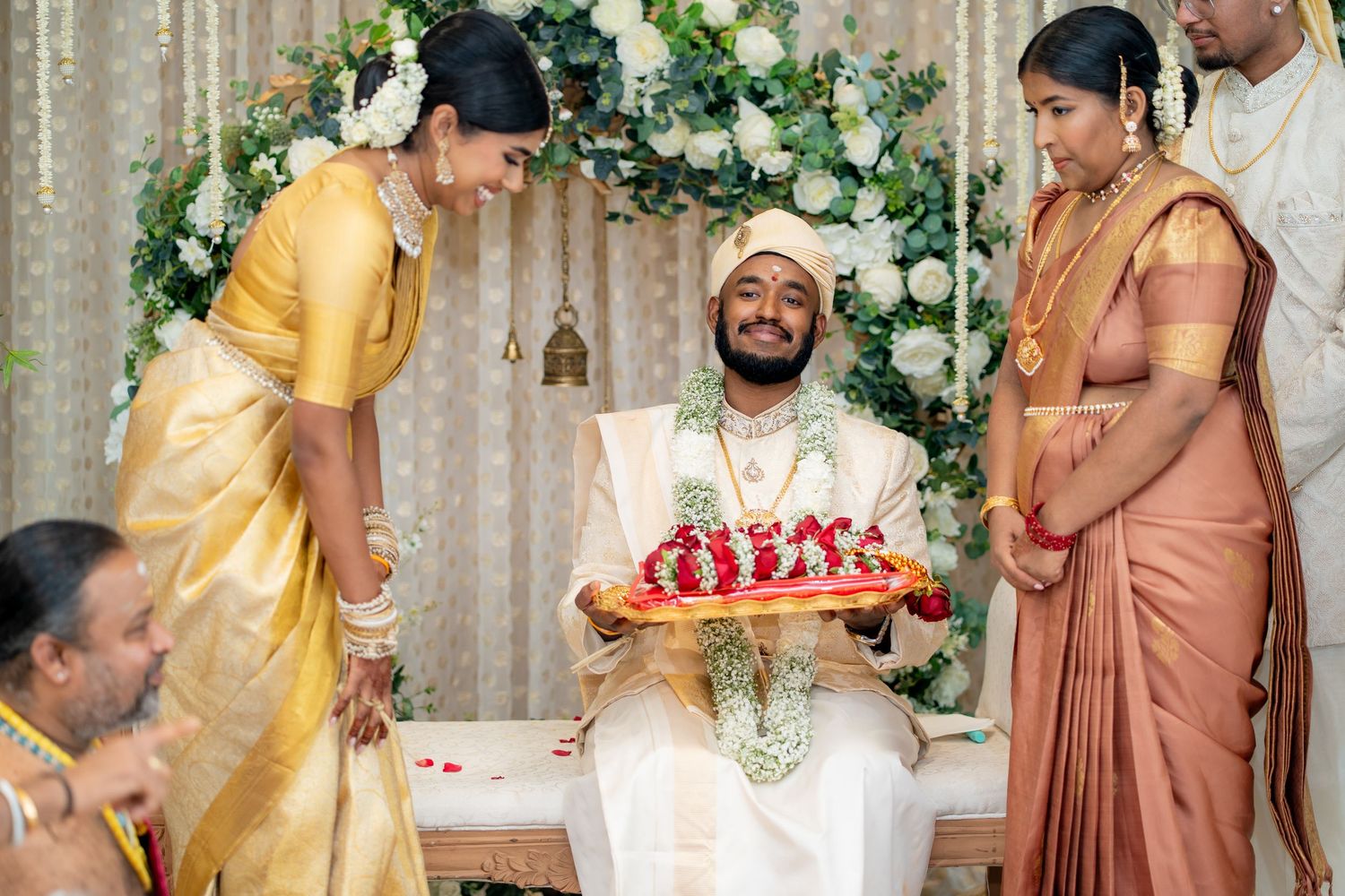 Traditional South Indian wedding ceremony with groom seated as bride presents floral tray, surrounded by family in silk attire.