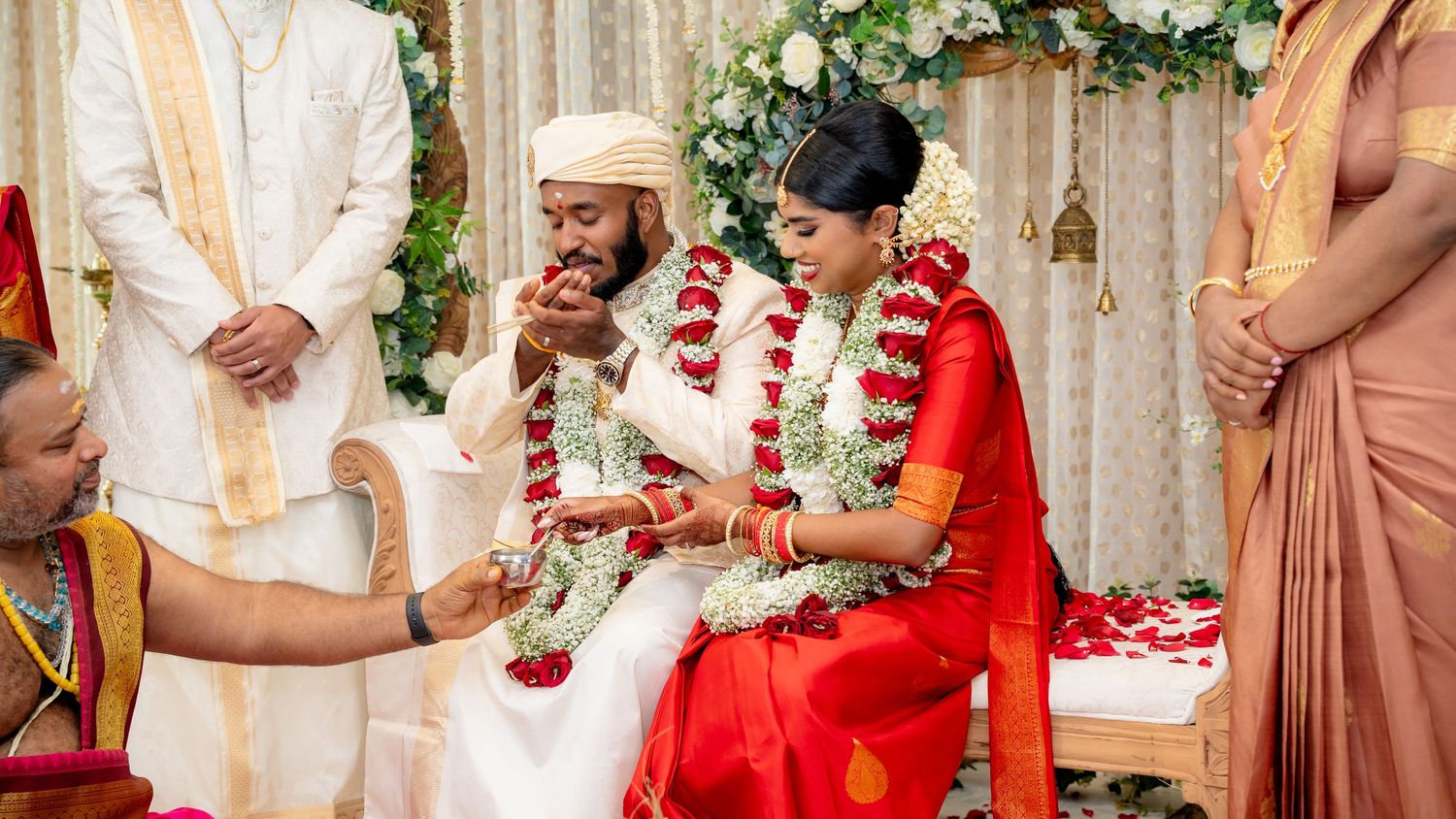 Smiling couple exchanges vows during colorful South Indian Hindu wedding ceremony with floral garlands.