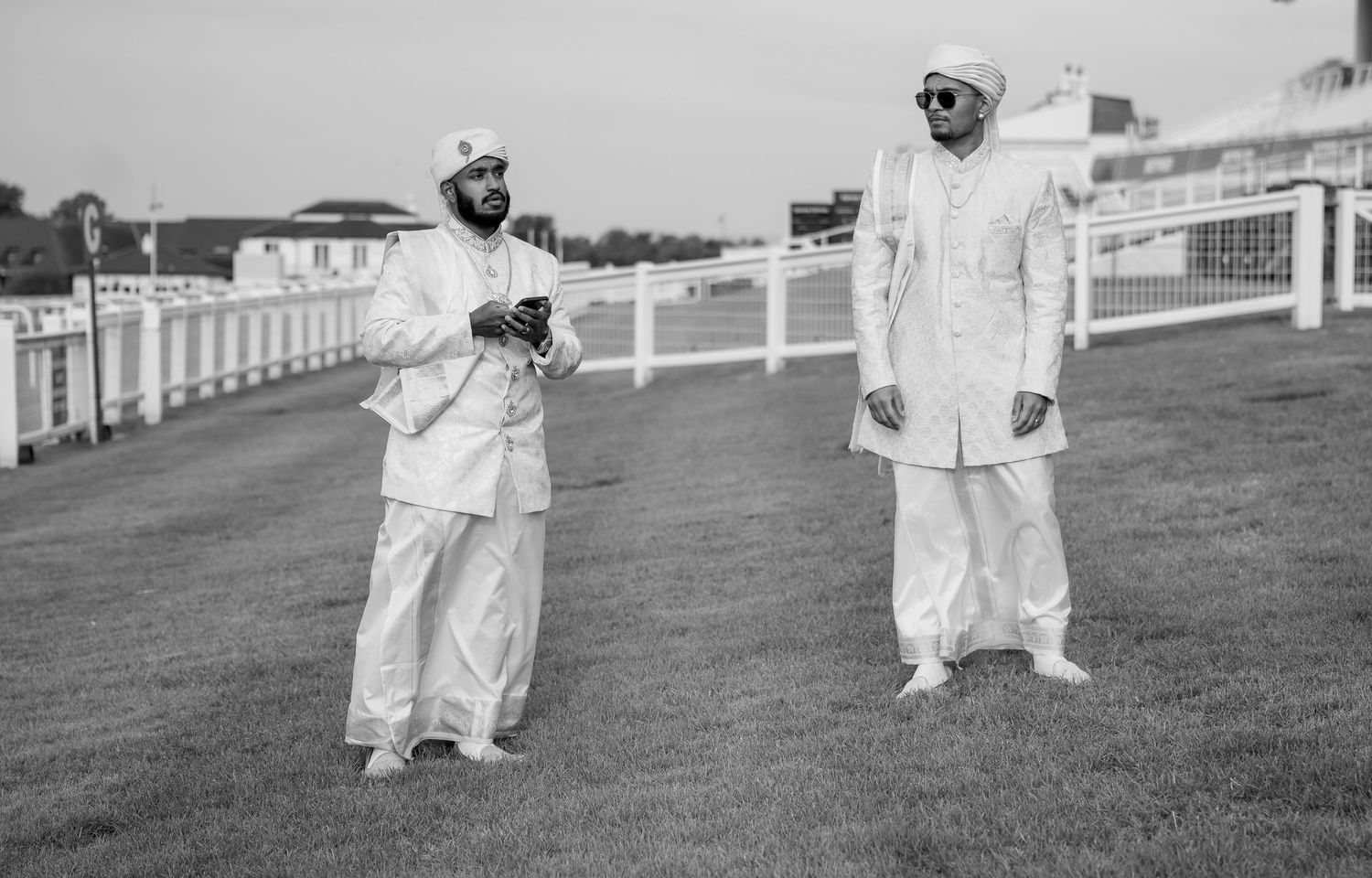 Two men in white traditional South Asian attire stand on a racecourse in this elegant black and white photo.