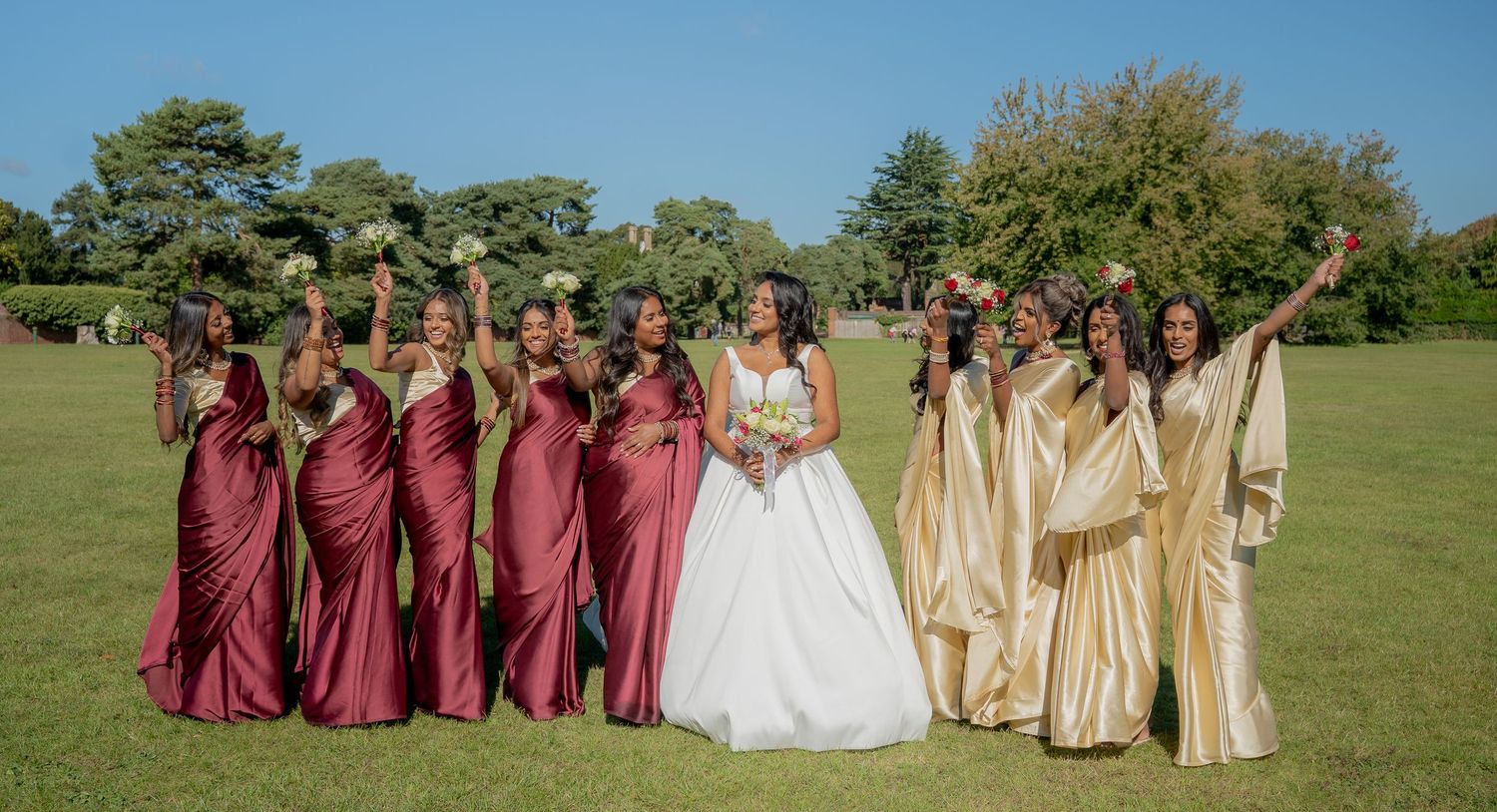 Bride and bridesmaids in burgundy and gold sarees celebrate joyfully on a sunny green lawn.