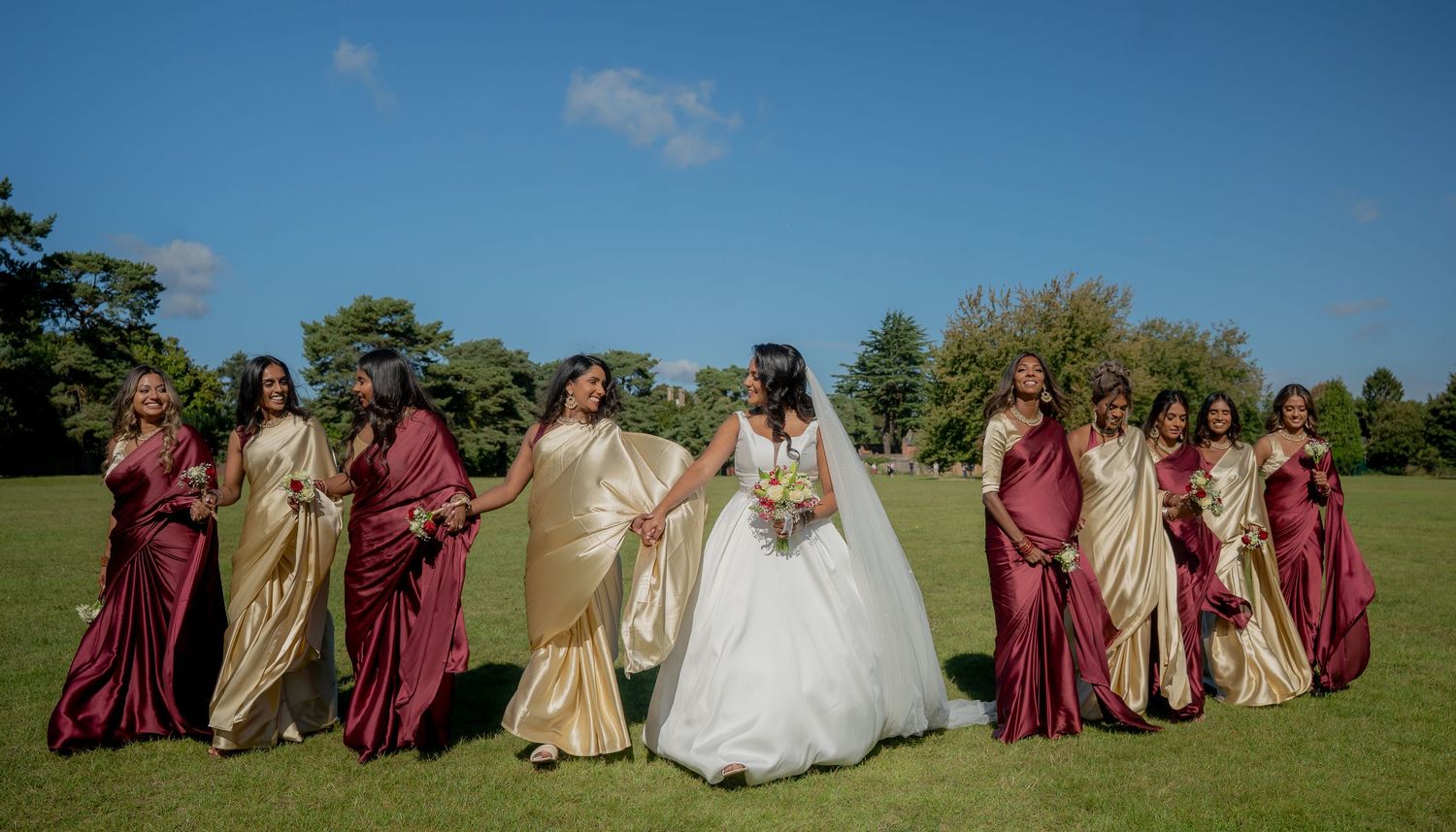 Bride in white ball gown holds hands with bridesmaids in burgundy and gold dresses on lush green lawn.