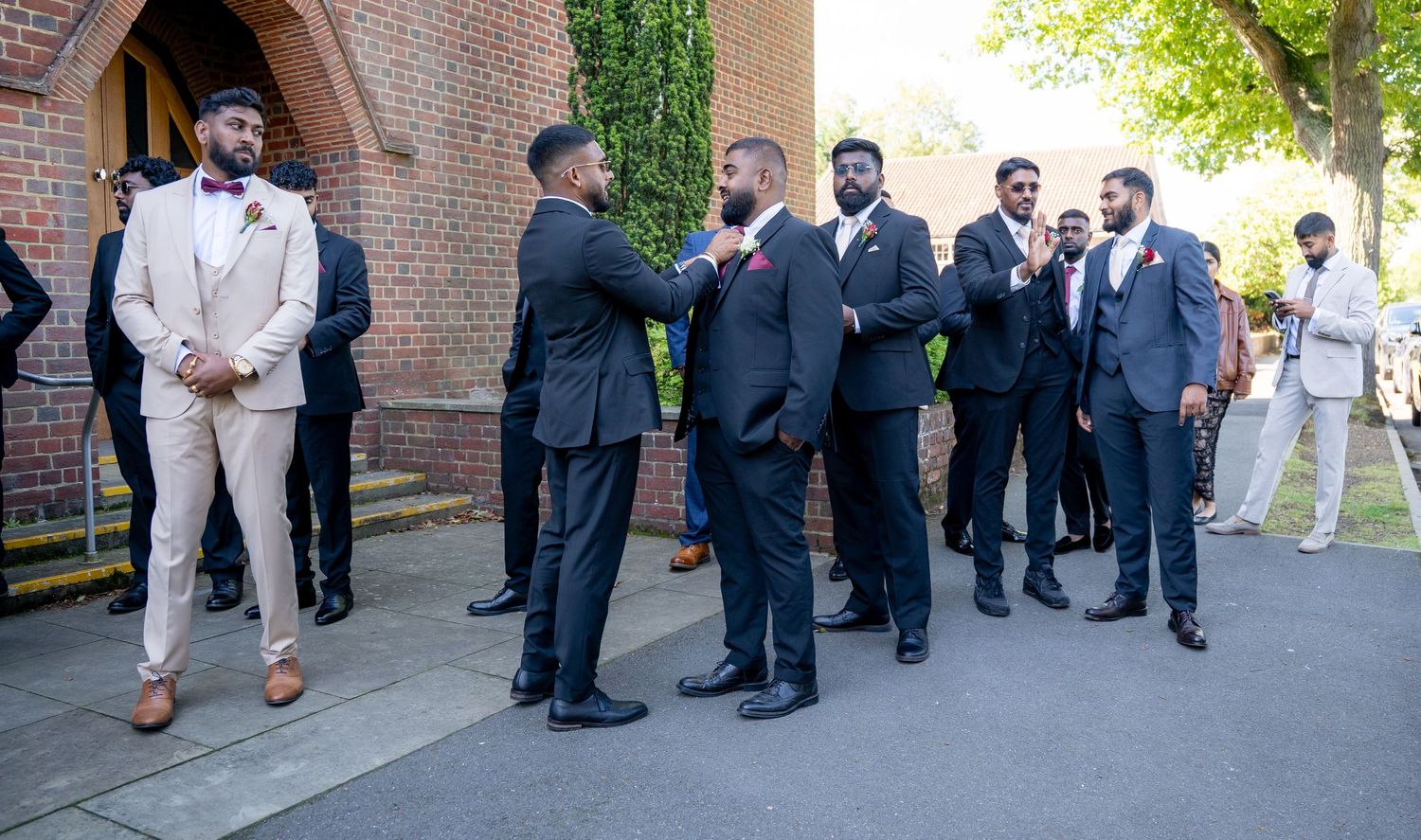 Groomsmen in navy suits adjusting ties outside church entrance on bright sunny wedding day.