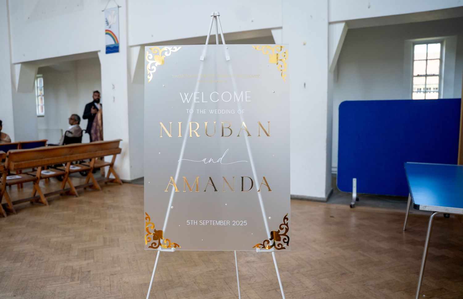 Elegant frosted acrylic wedding welcome sign for Niruban and Amanda on a gold easel in a church hall.