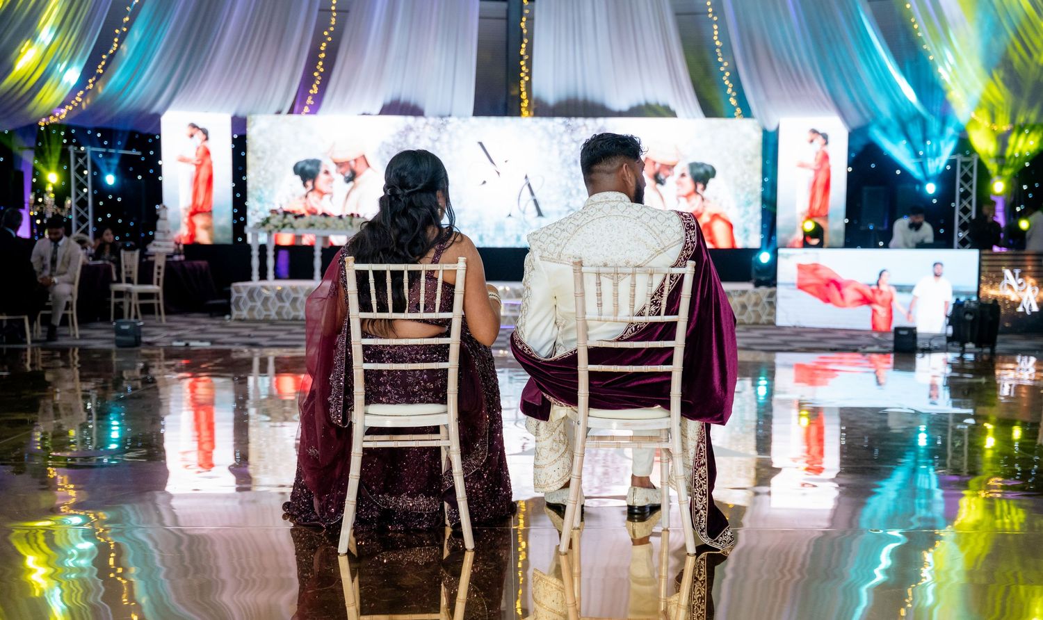 Bride and groom sit together on dance floor watching their wedding photos on a large display screen.