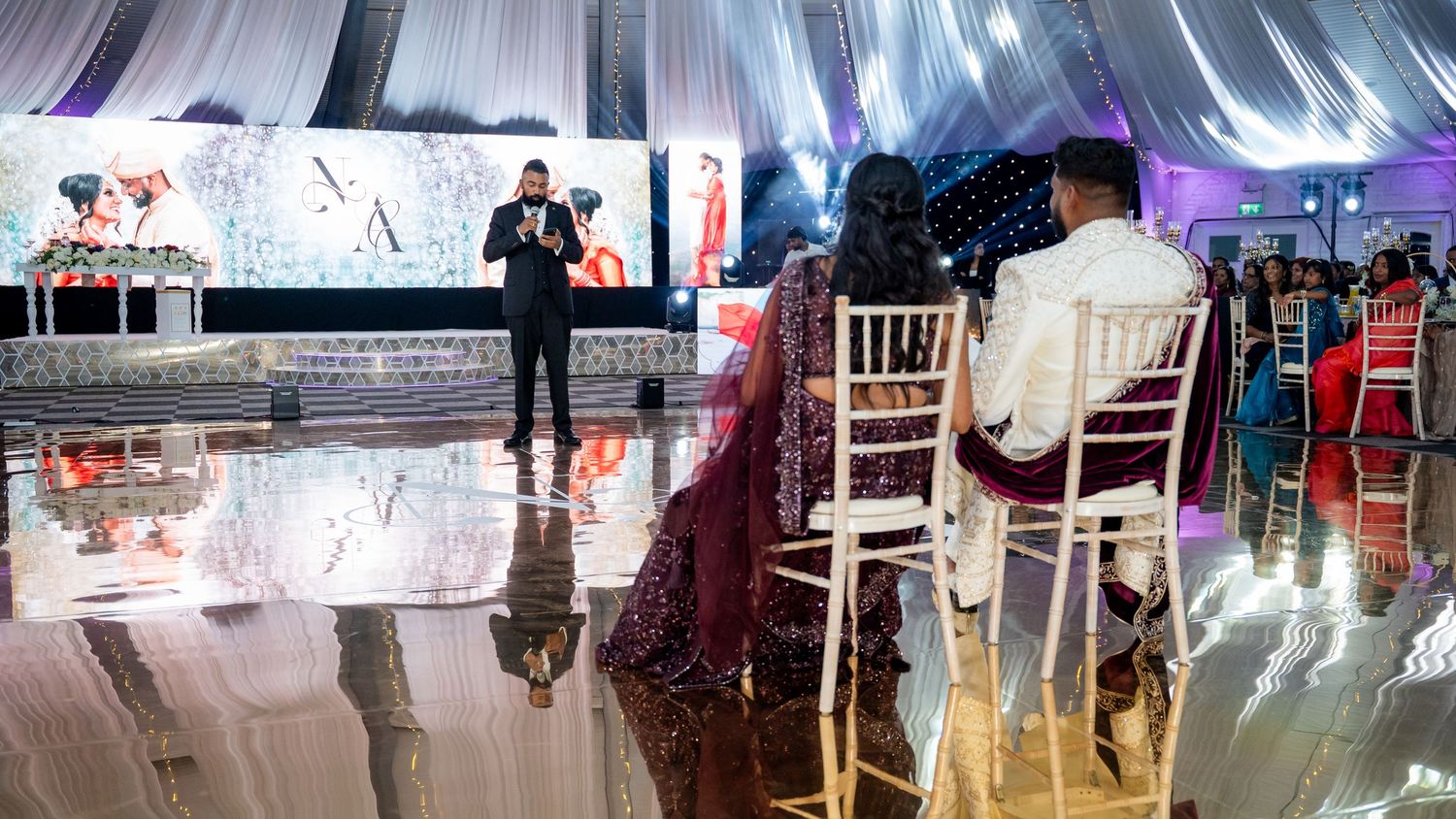 Elegant wedding reception with couple seated on chairs while MC speaks on a decorated dance floor.