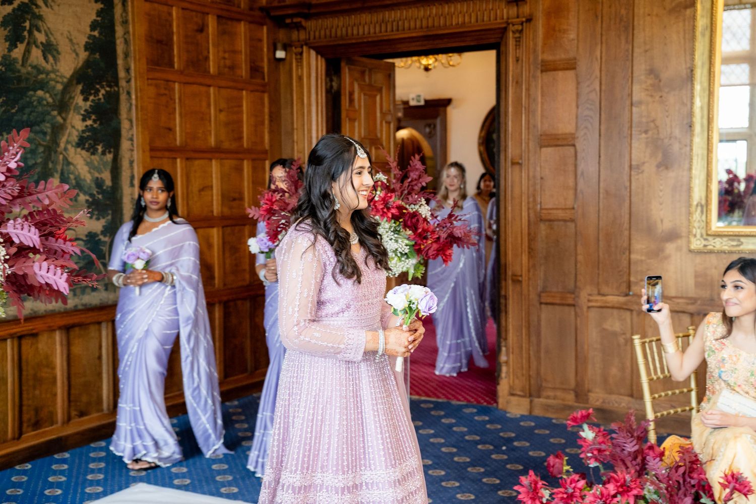 Bride in pink lehenga walks through ornate wooden doorway holding red floral bouquet with bridesmaids in blue sarees.