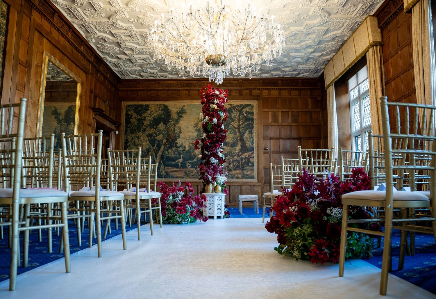 Elegant wedding ceremony room with gold chiavari chairs, chandelier, and red floral arrangements.
