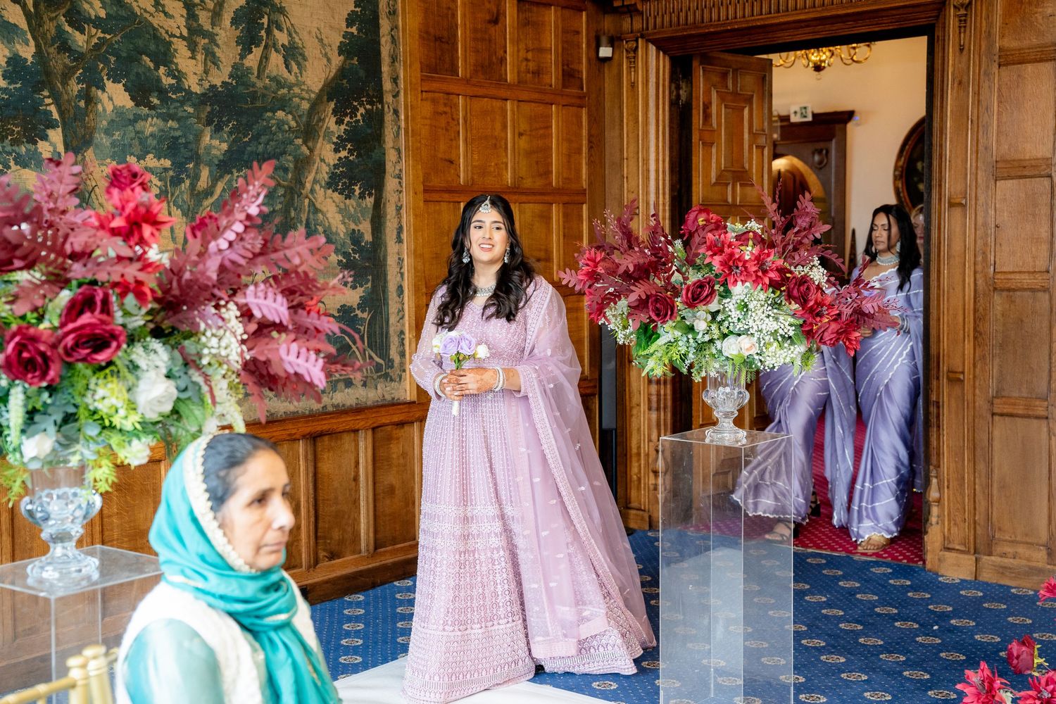 Bride in pink lehenga stands at ornate doorway with red floral arrangements at Indian wedding ceremony.