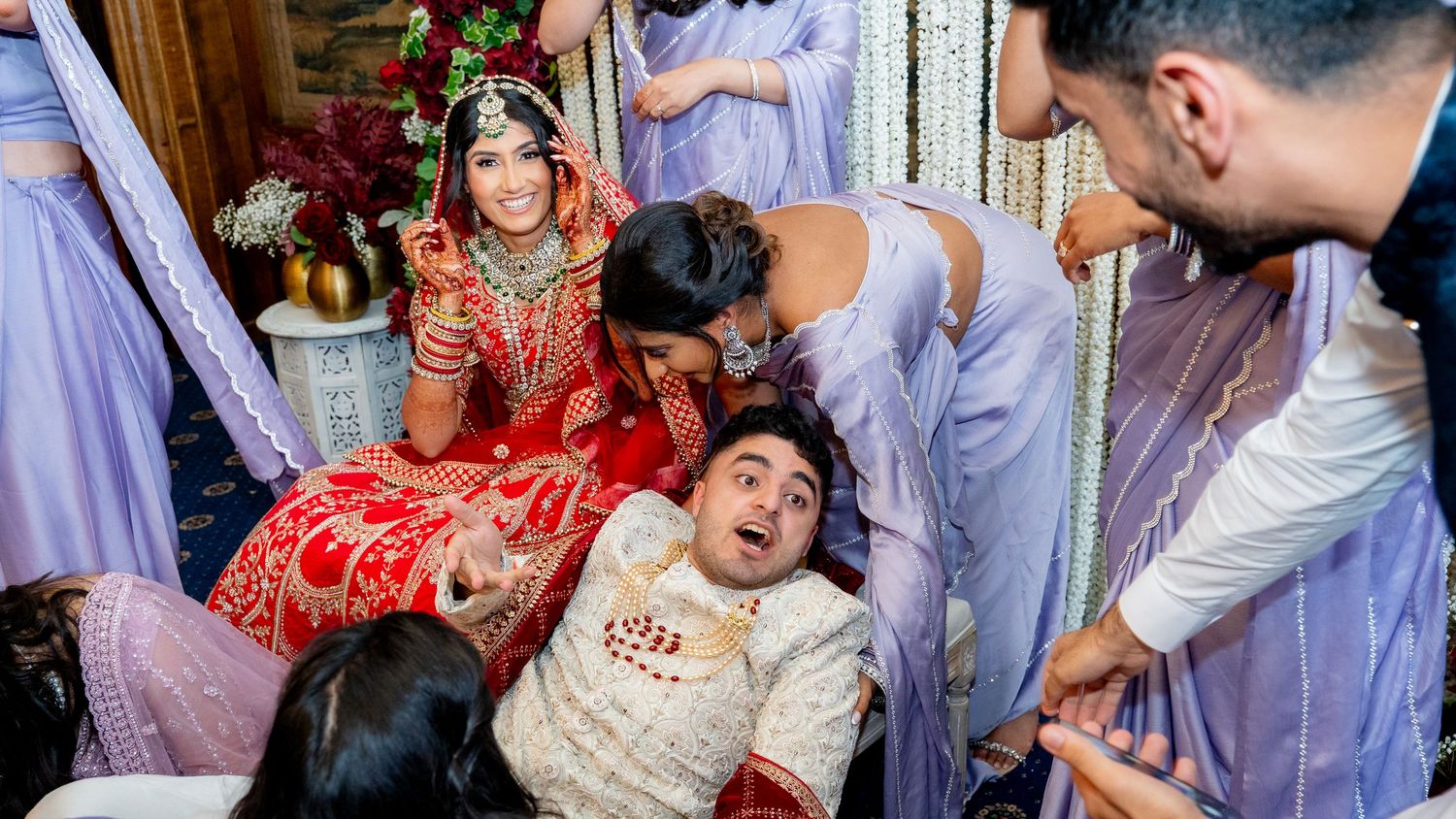 Bride in red bridal lehenga surrounded by laughing bridesmaids during fun wedding celebration moment.