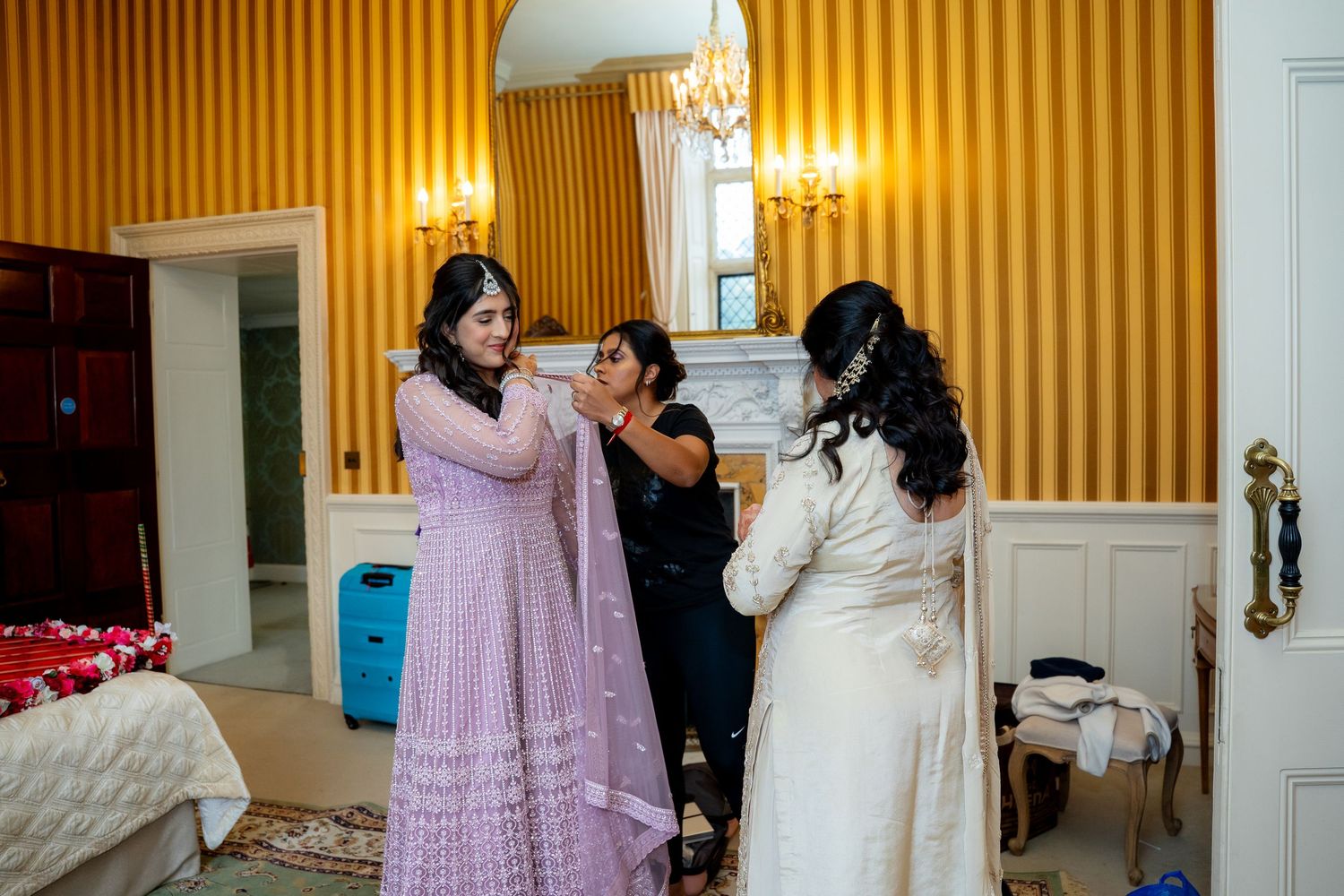 Women helping bride dress in yellow-walled room, wearing Indian attire during traditional wedding preparation.