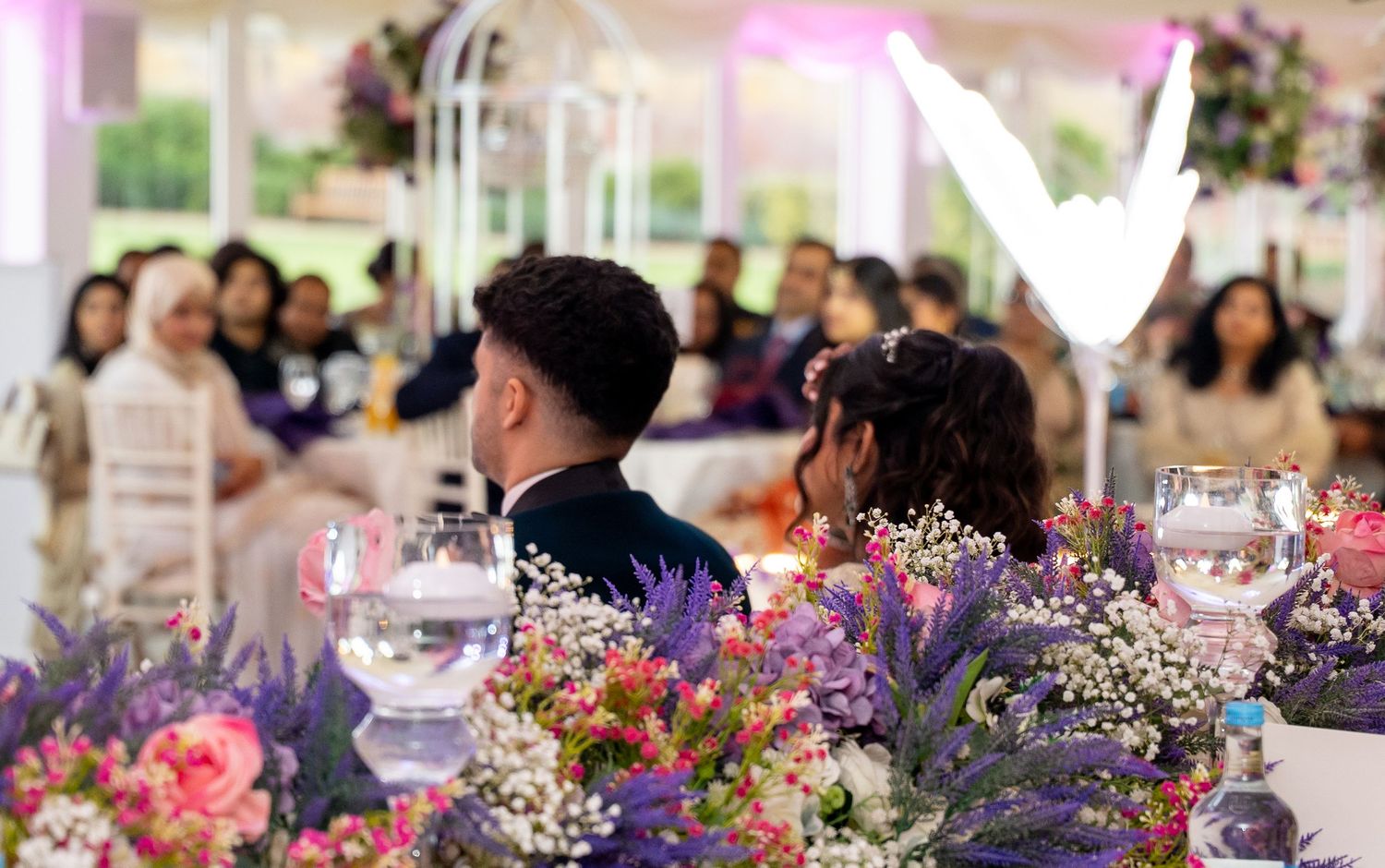 Wedding couple seated at floral decorated head table with colorful pink and purple flower arrangements.