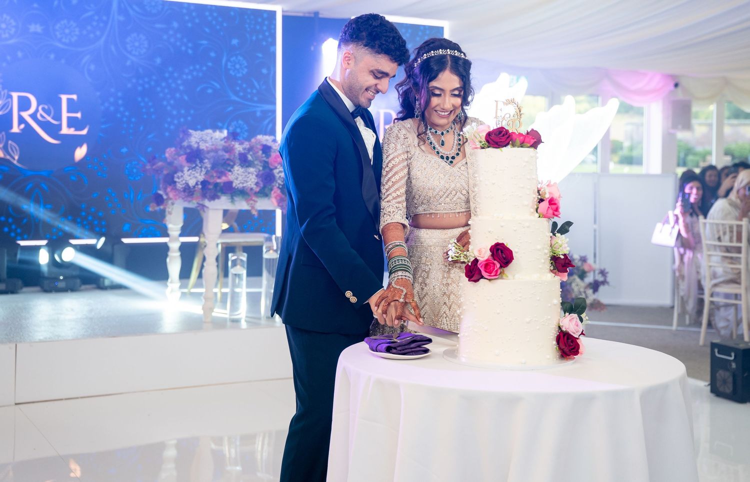 Couple in wedding attire cutting elegant white three-tier cake decorated with red roses at reception.