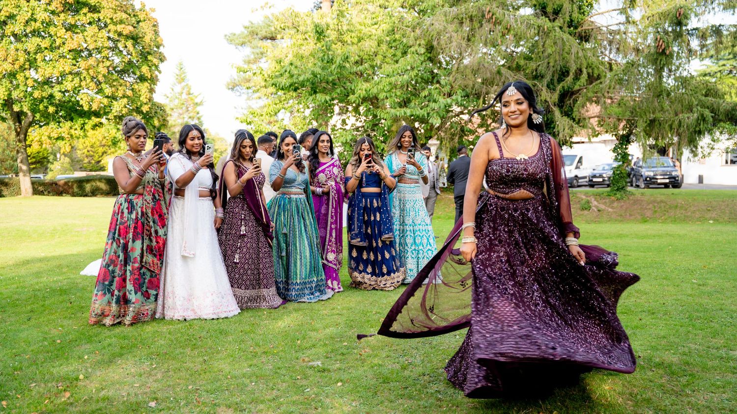 A bride twirls her purple lehenga on green lawn while bridesmaids in colorful Indian attire cheer behind her.