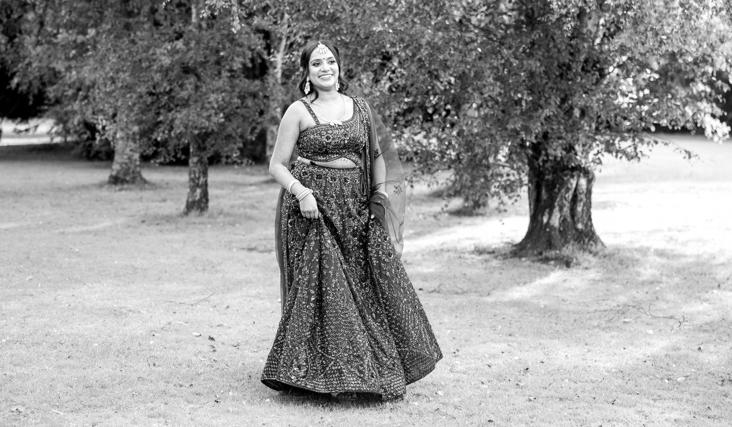 Black and white photo of woman in embroidered lehenga posing in a lush park setting.
