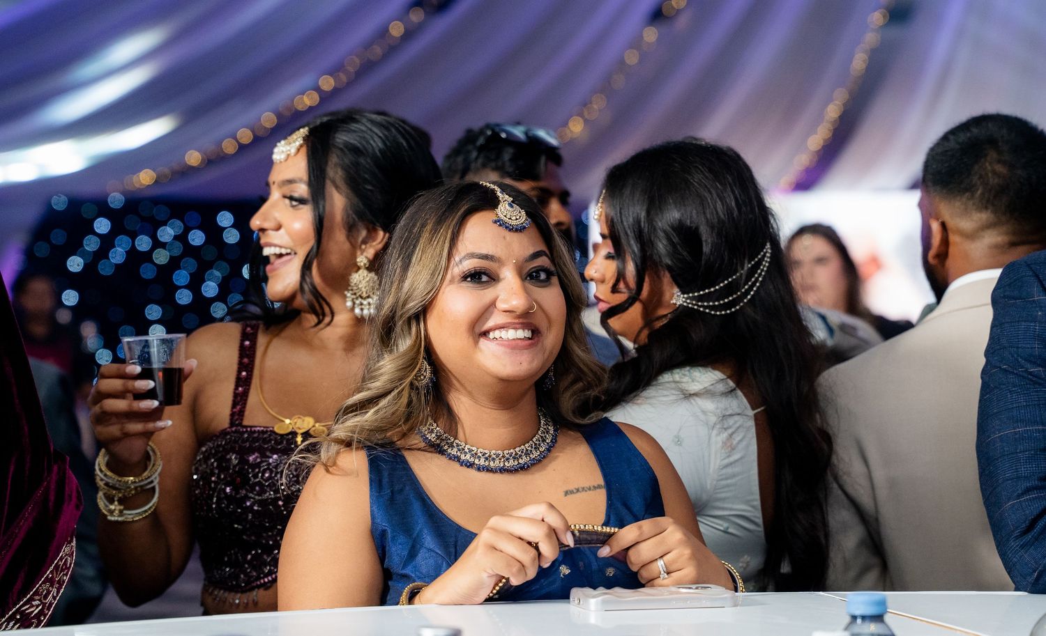 Smiling woman in blue lehenga with maang tikka enjoys Indian wedding reception with friends behind her.