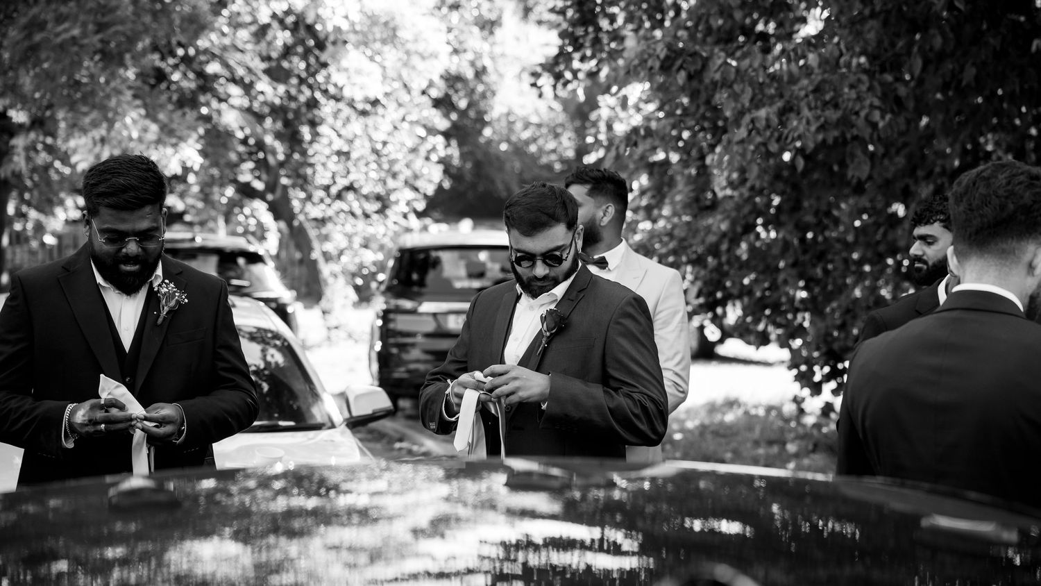 Black and white photo of groomsmen adjusting boutonnieres beside cars under tree canopy.