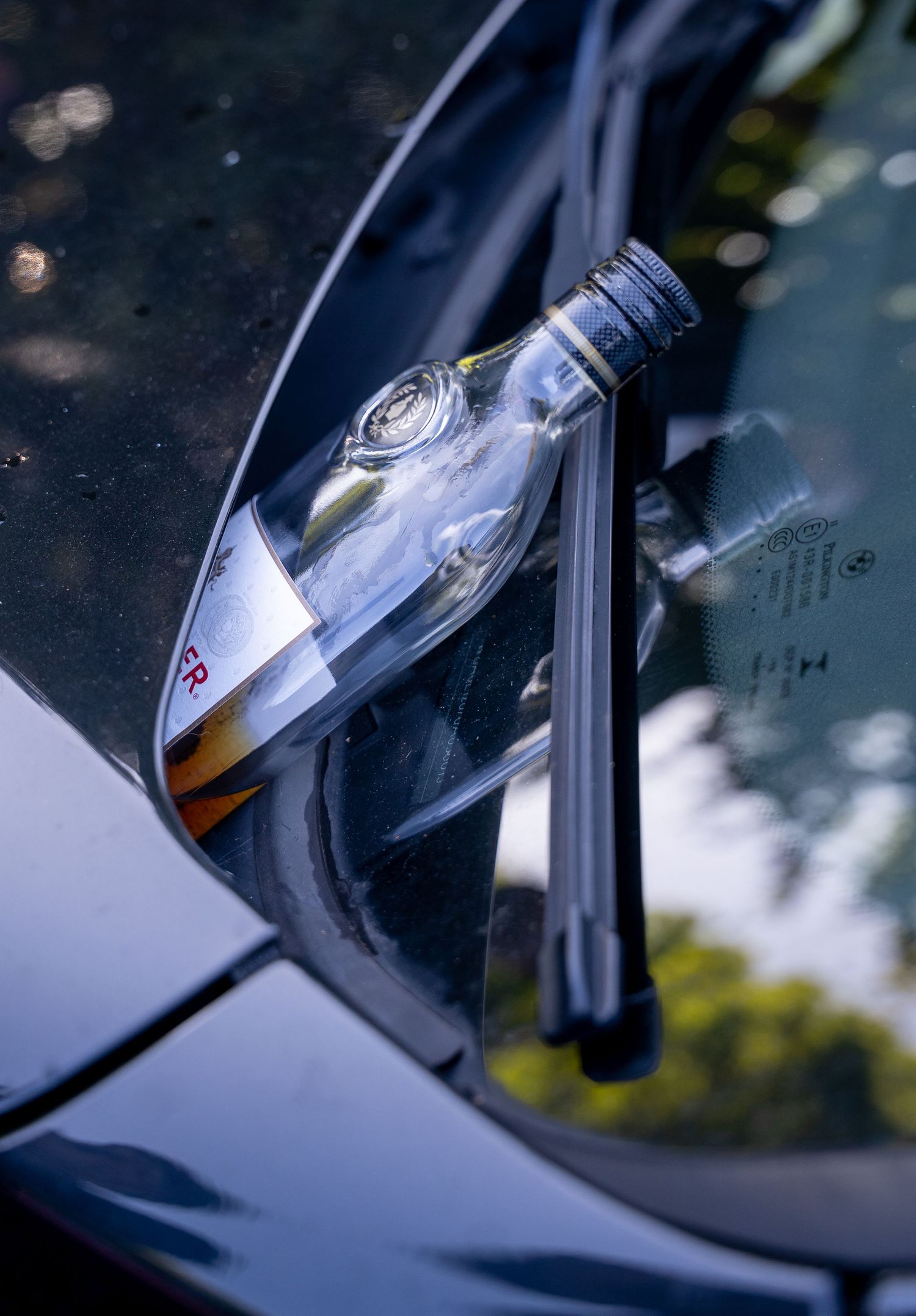 Liquor bottle wedged under car windshield wiper on vehicle hood outdoors.