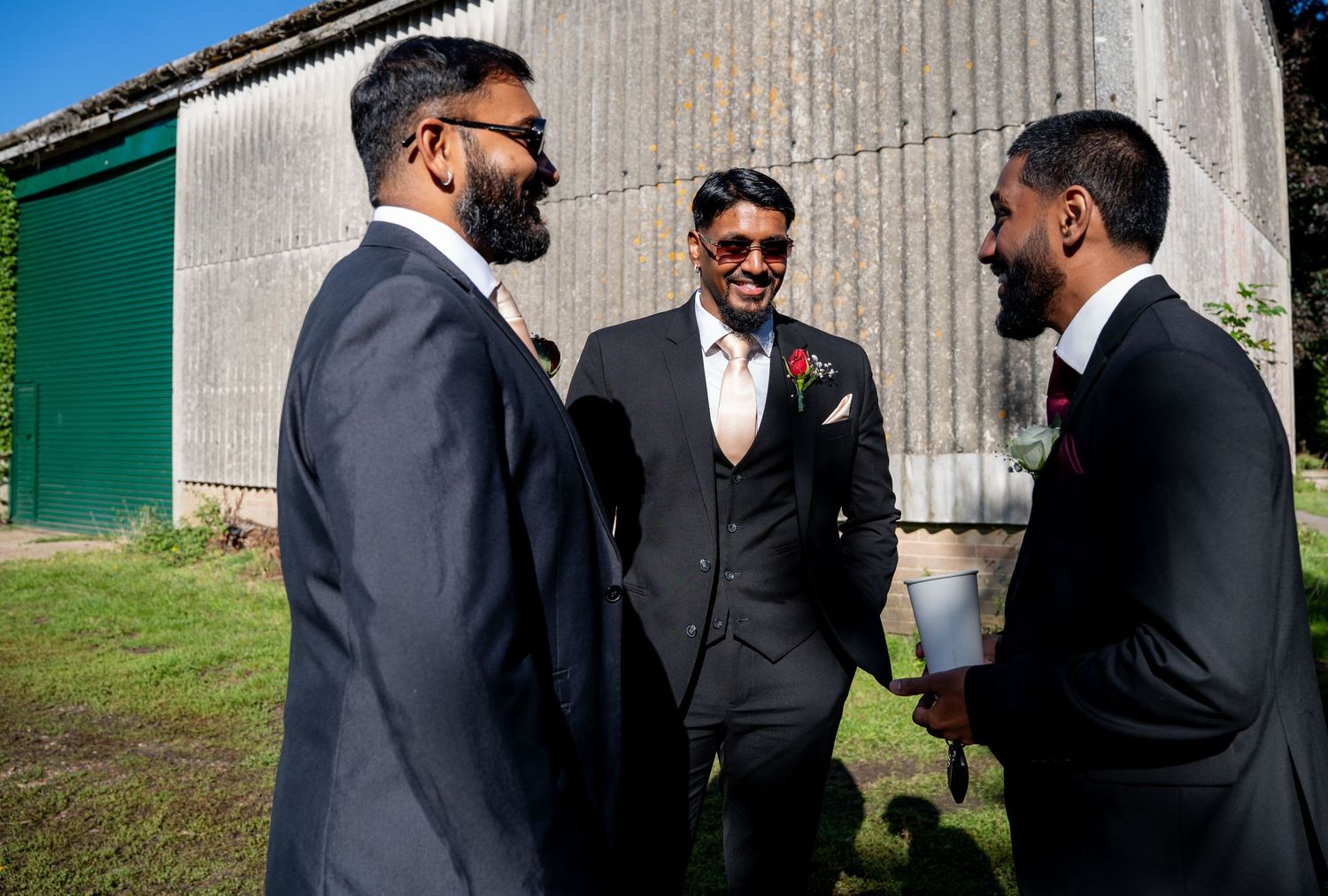 Three groomsmen in dark suits laugh together outside a rustic barn on wedding day.