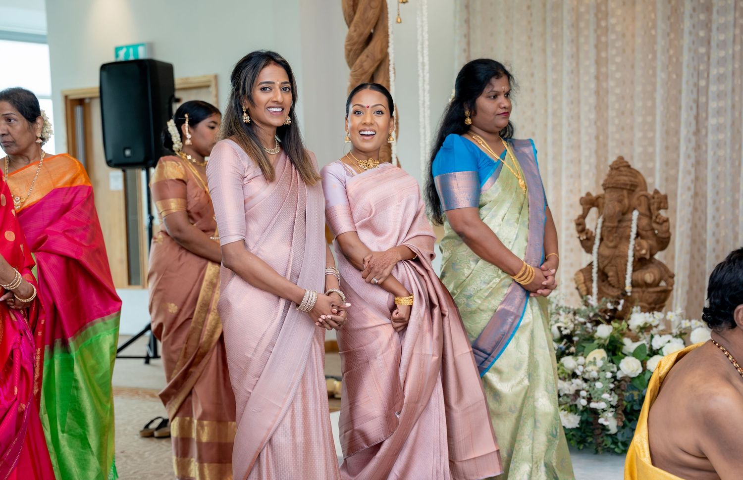 Two women in pink sarees smile at an Indian wedding celebration with guests in colorful traditional attire.