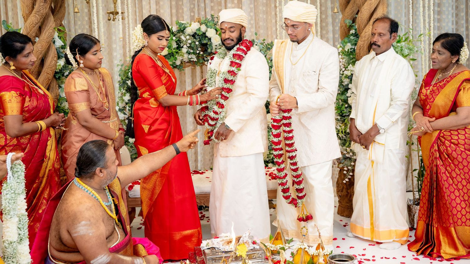 South Indian wedding ceremony with bride in red saree and groom in white sherwani during traditional garland exchange.