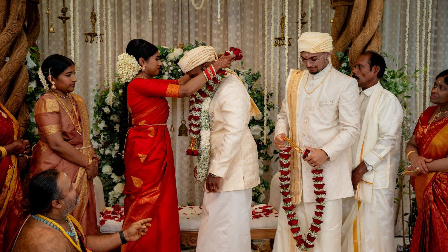 Traditional South Indian bride in red saree places floral garland on groom in white sherwani during wedding ritual.