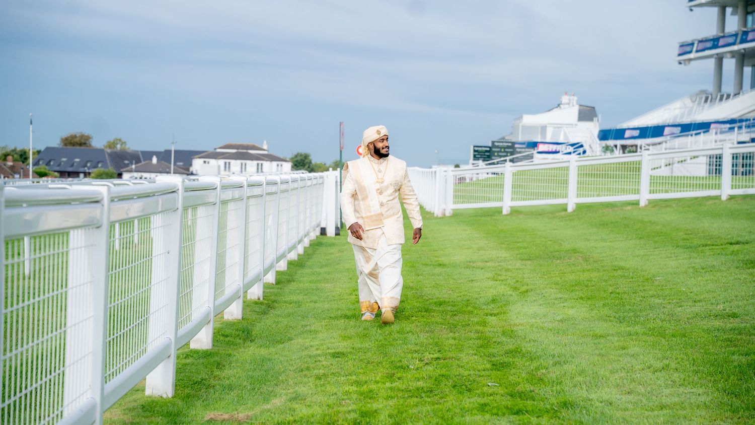 A man in traditional Arab white thobe walks along a green grass racecourse with white fencing on a cloudy day.
