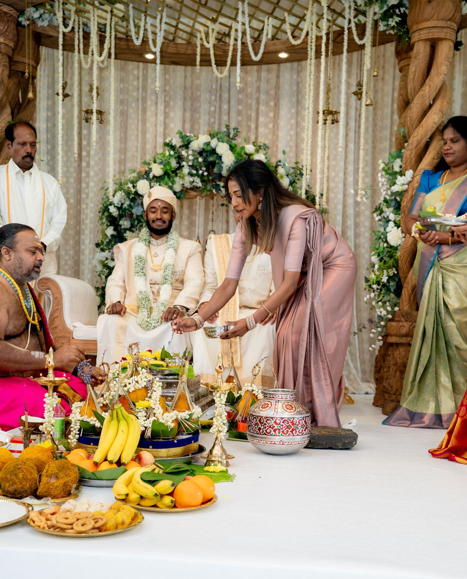Bride in pink saree performs wedding rituals at a decorated Hindu mandap with offerings and flowers.