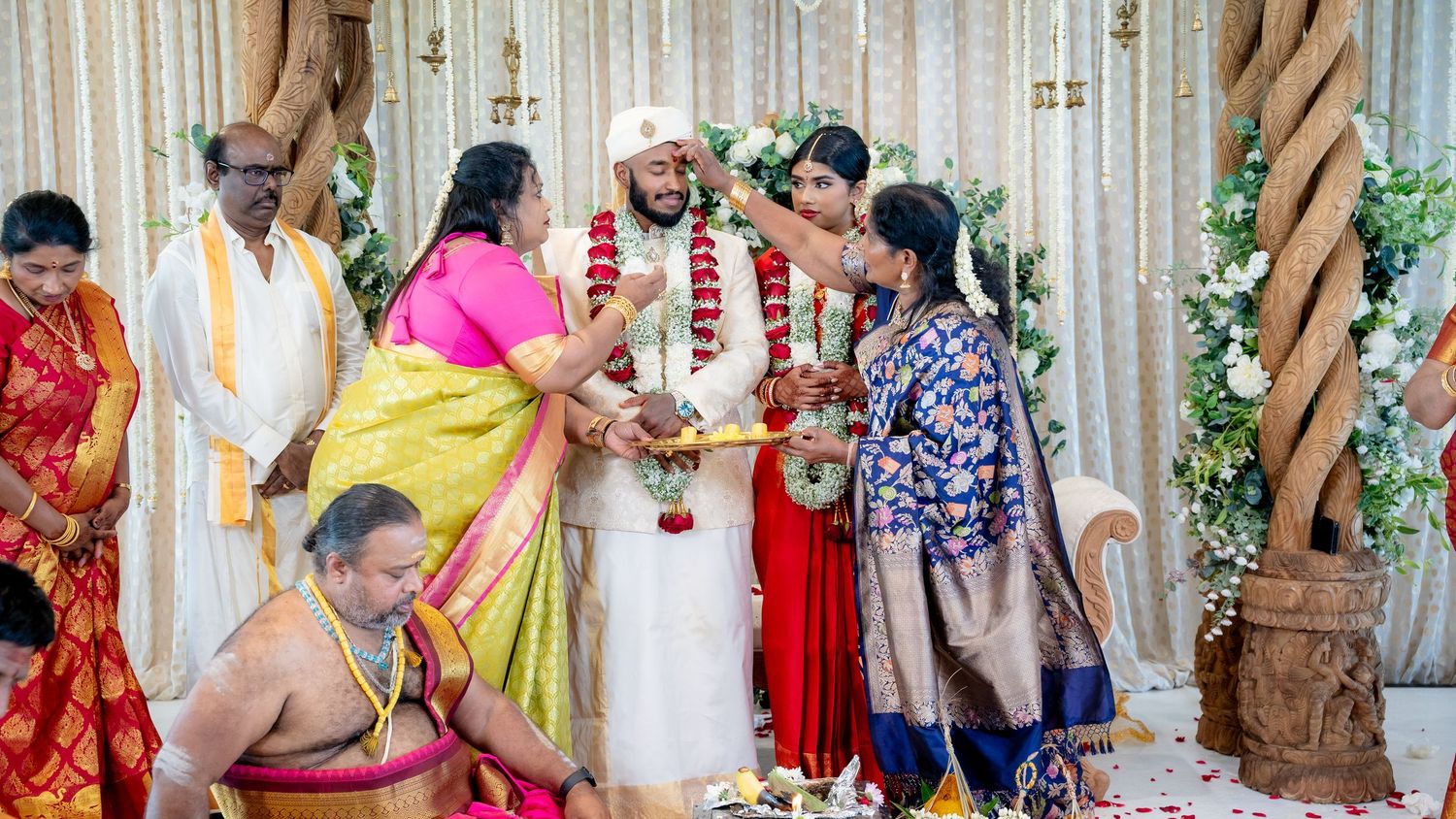 South Indian wedding garland exchange ceremony with priest and family members in colorful silk sarees on floral mandap.