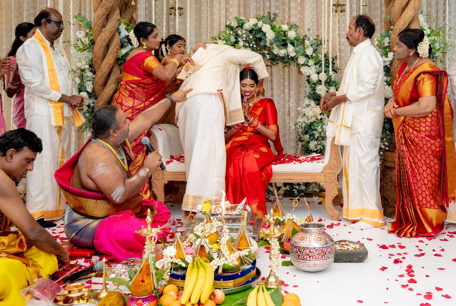South Indian wedding ritual with groom bowing to seated bride in red saree surrounded by family and priest.