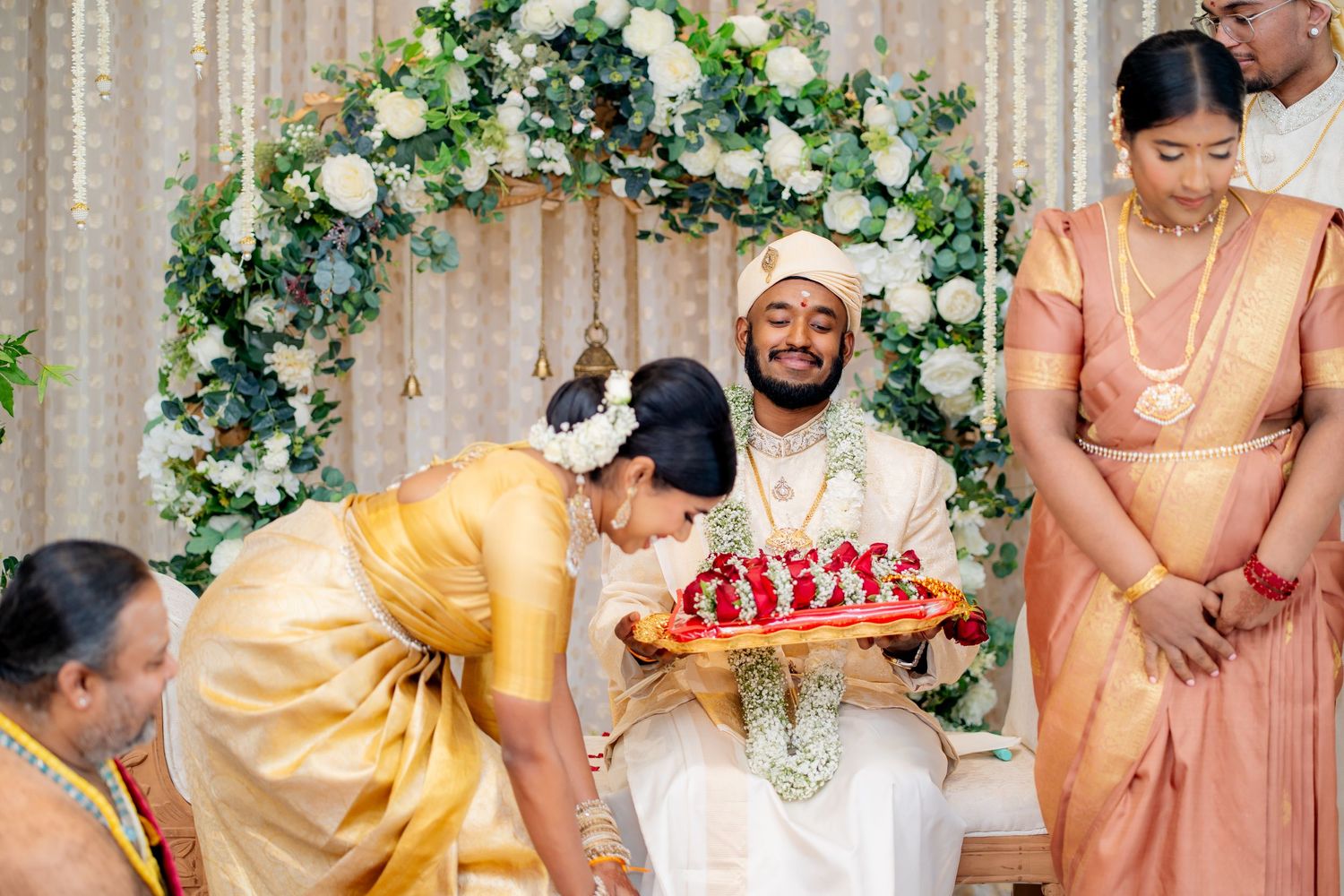 Traditional wedding ceremony with groom seated, bride in yellow attire, and floral arch backdrop with white and green flowers.