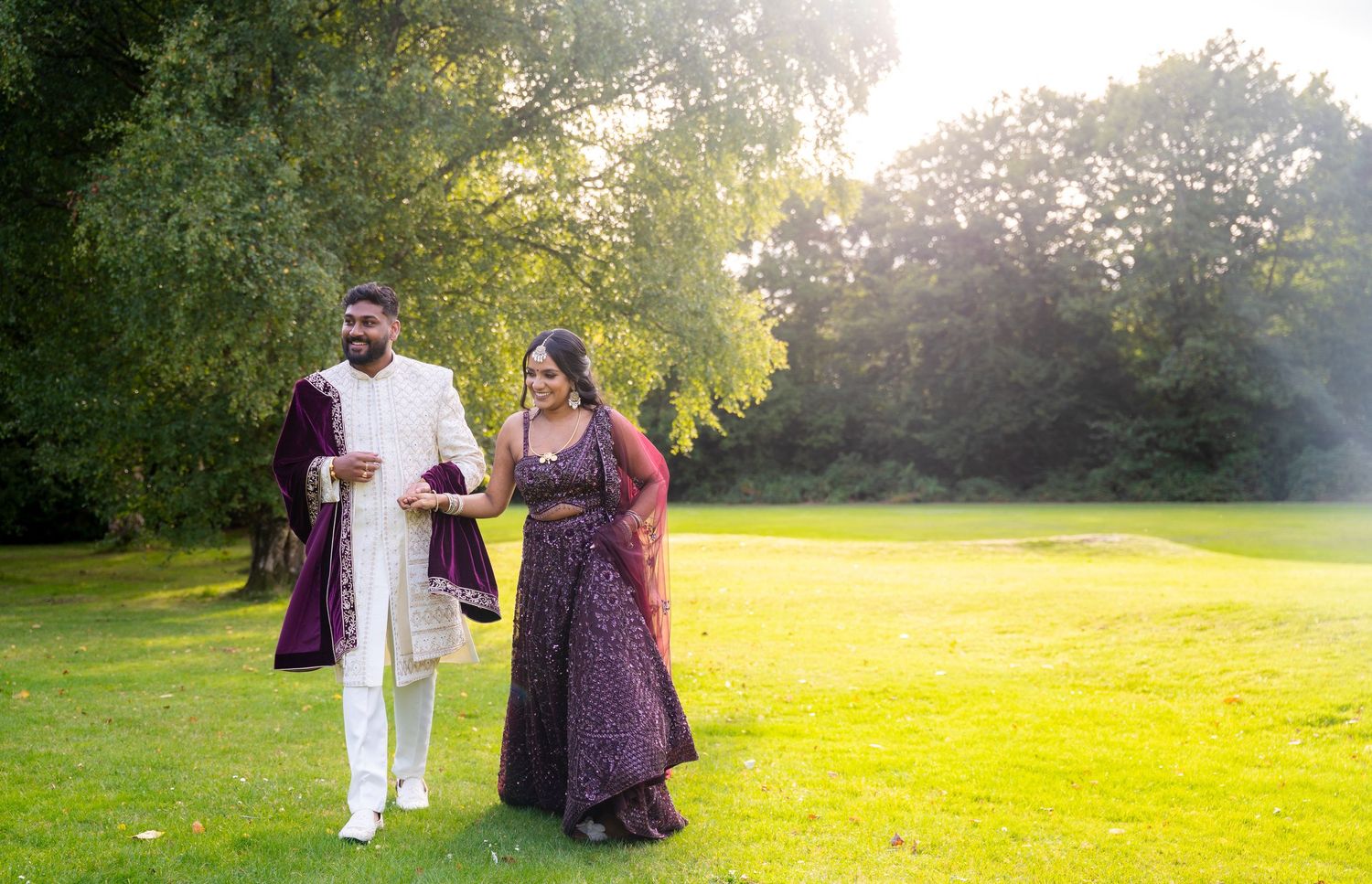 Indian bride and groom in traditional wedding attire strolling through sunlit green park grounds.