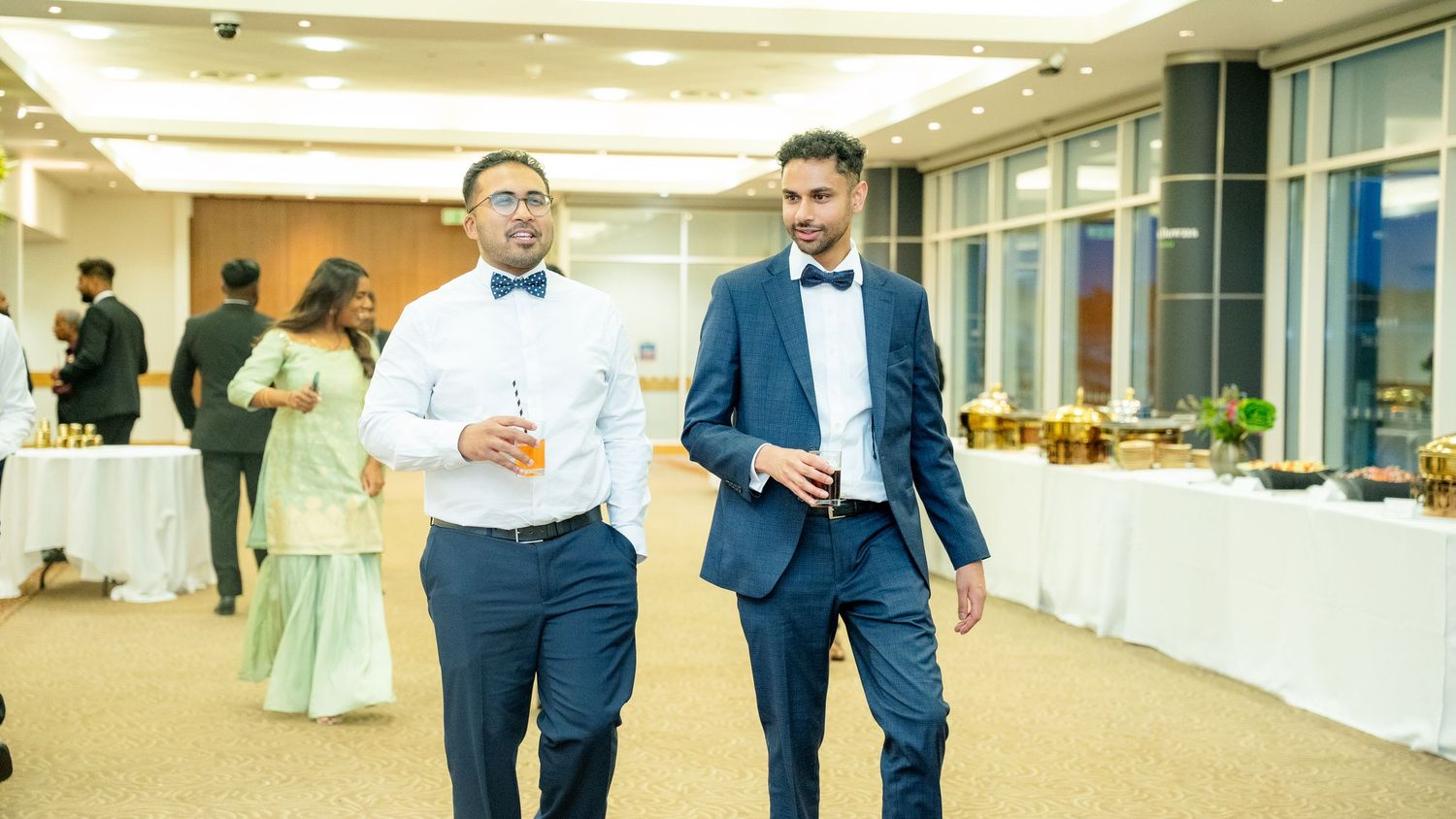 Two men in suits with bow ties conversing while walking through an elegant event venue.
