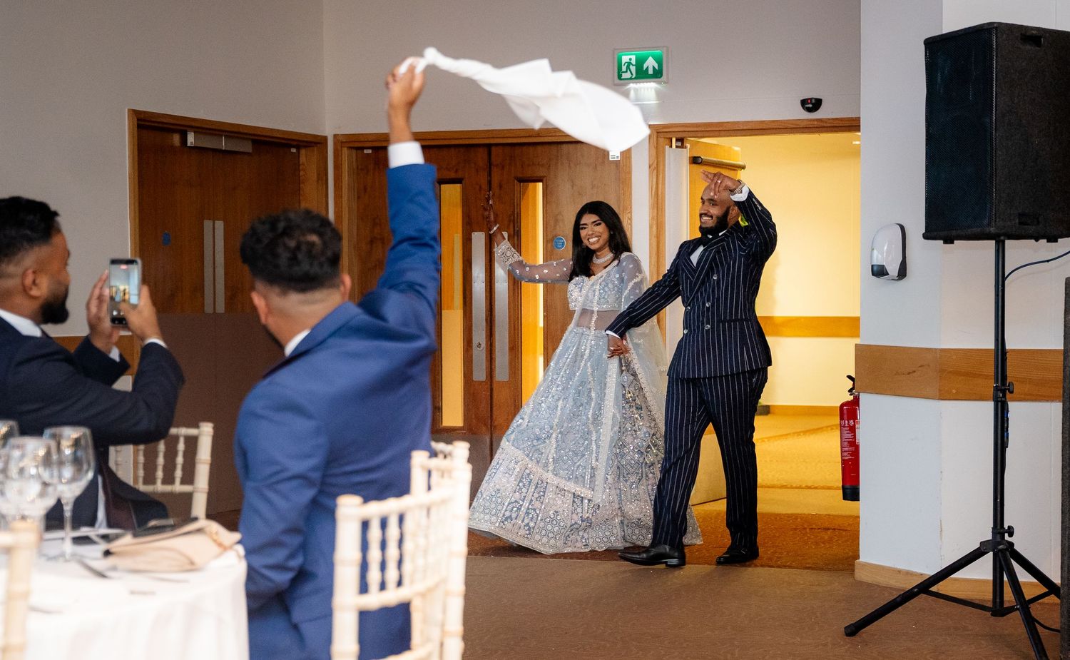 Bride in blue lehenga and groom make grand entrance as guest waves white cloth at wedding reception.