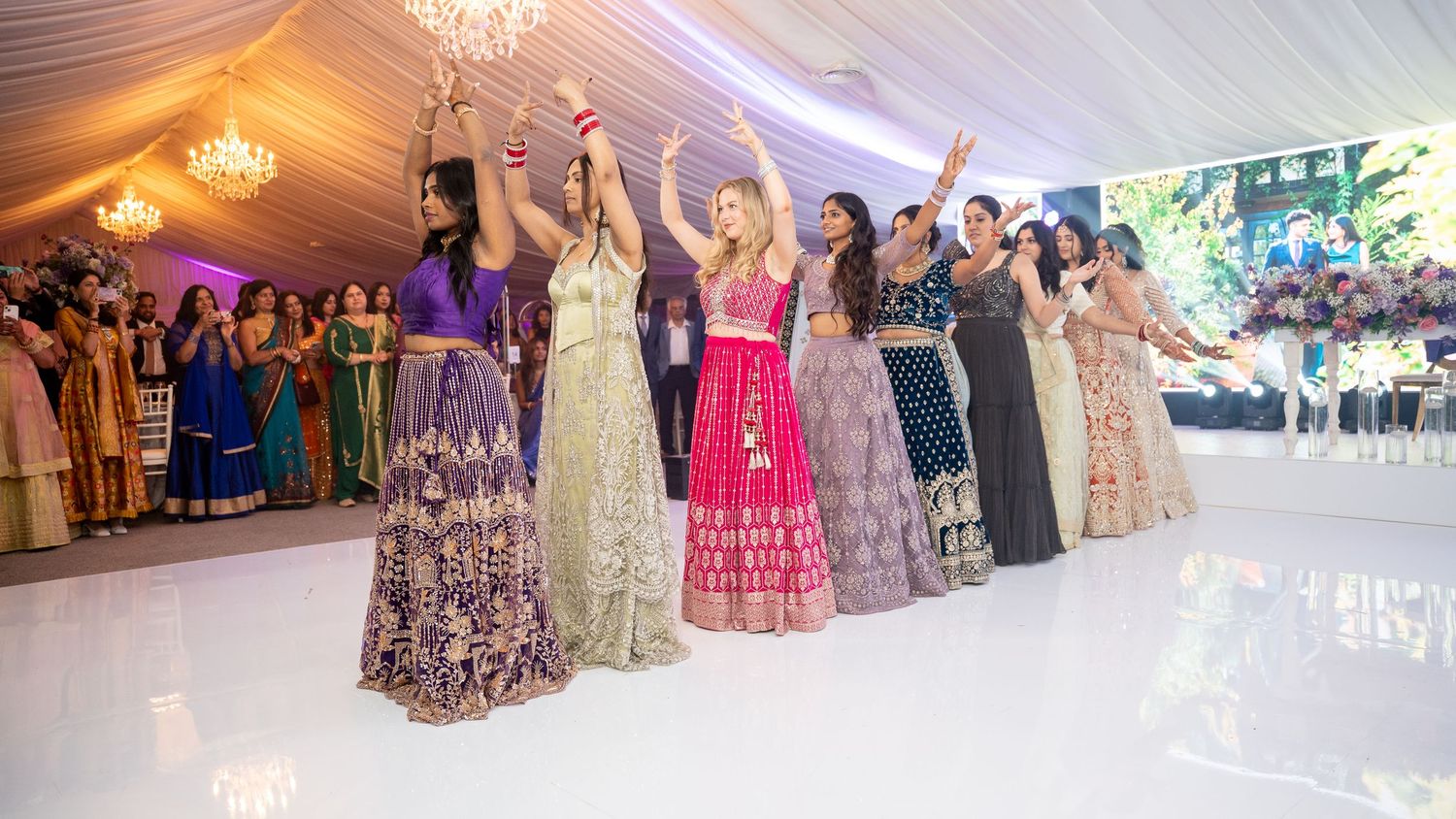 Group of women in vibrant Indian attire dancing joyfully at a wedding reception under chandeliers.