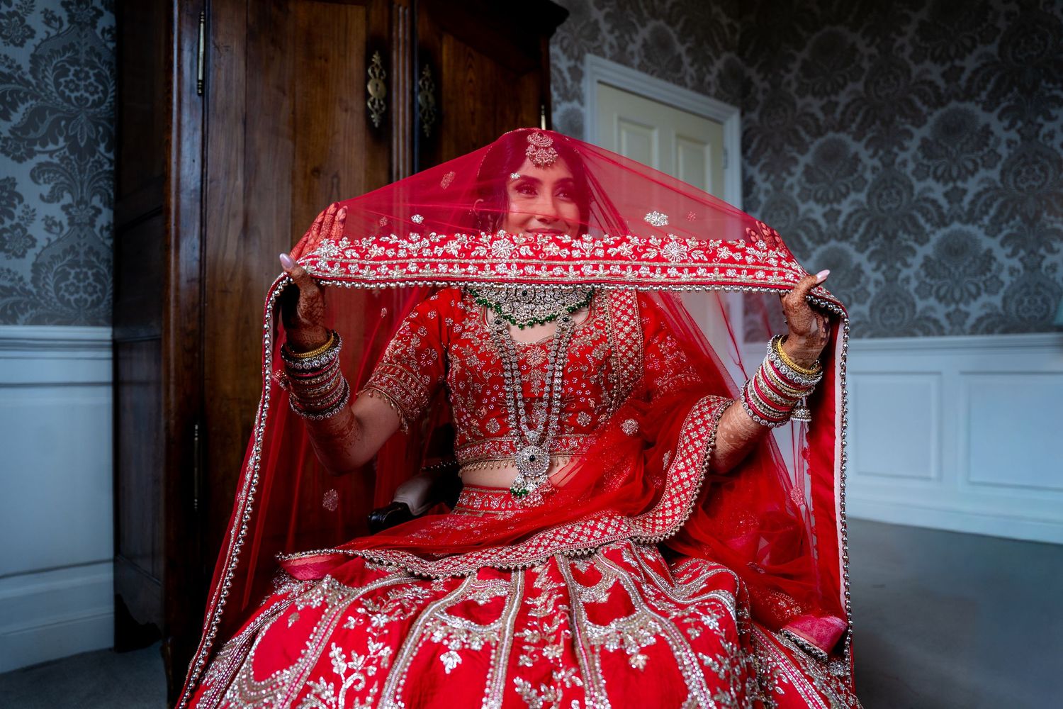 Indian bride in red lehenga holds embroidered dupatta overhead in ornate blue room.